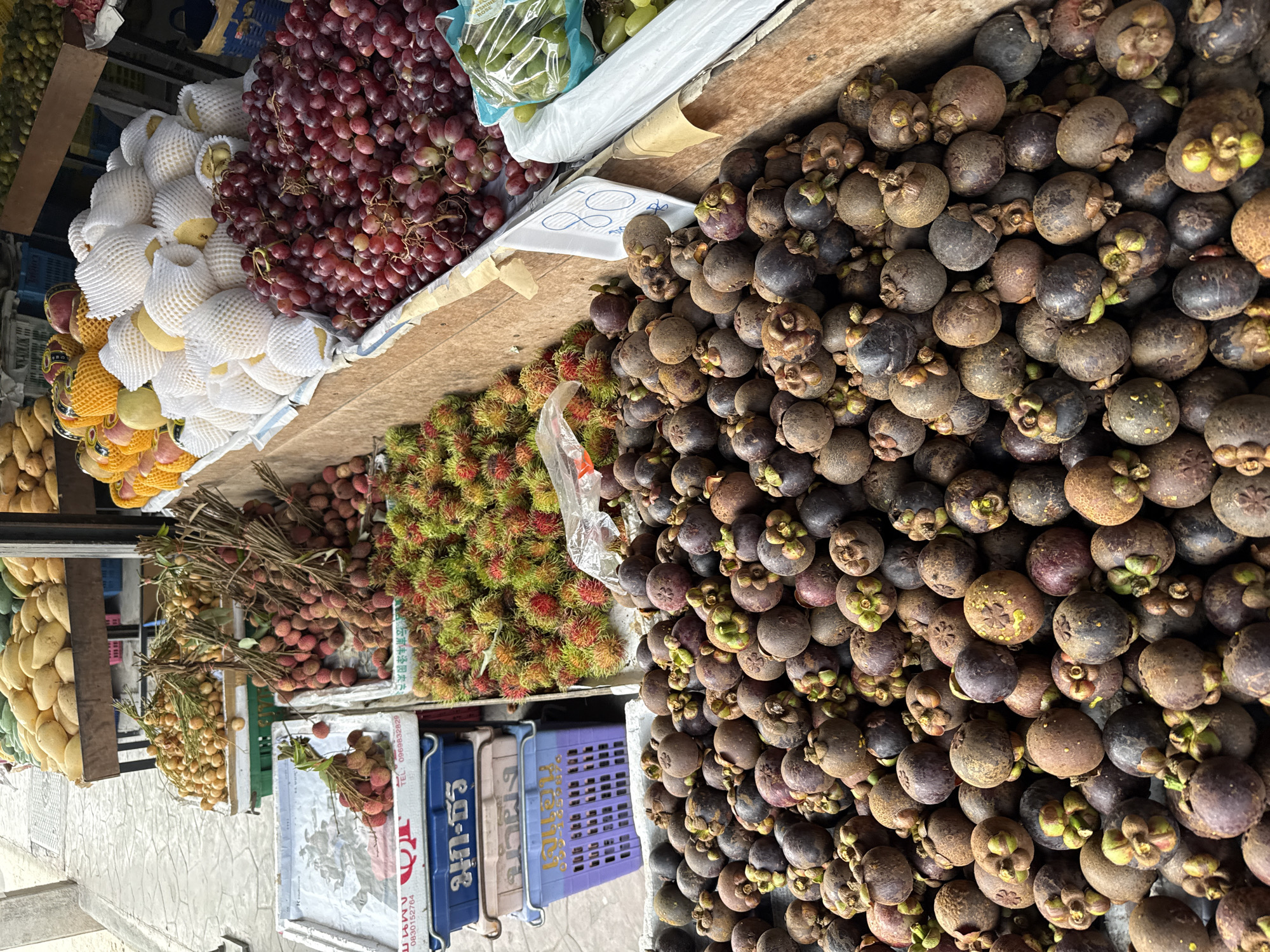 The image shows a street-side fruit stall in Moo 1, Thailand. A variety of fruits are displayed, including mangosteens, grapes, pears encased in white protective netting, rambutans, and lychees. The mangosteens dominate the right side of the image, forming a dense pile. Adjacent to them, on the left, are clusters of rambutan, their characteristic red and green spiky skins visible. Further to the left are bunches of lychees, their red skins and clustered arrangement apparent. On the leftmost side, pears are seen wrapped in white protective netting. Grapes, deep red in color, are also present alongside the pears. The fruits are arranged on a table or platform. Below the fruit, blue plastic crates are partially visible.