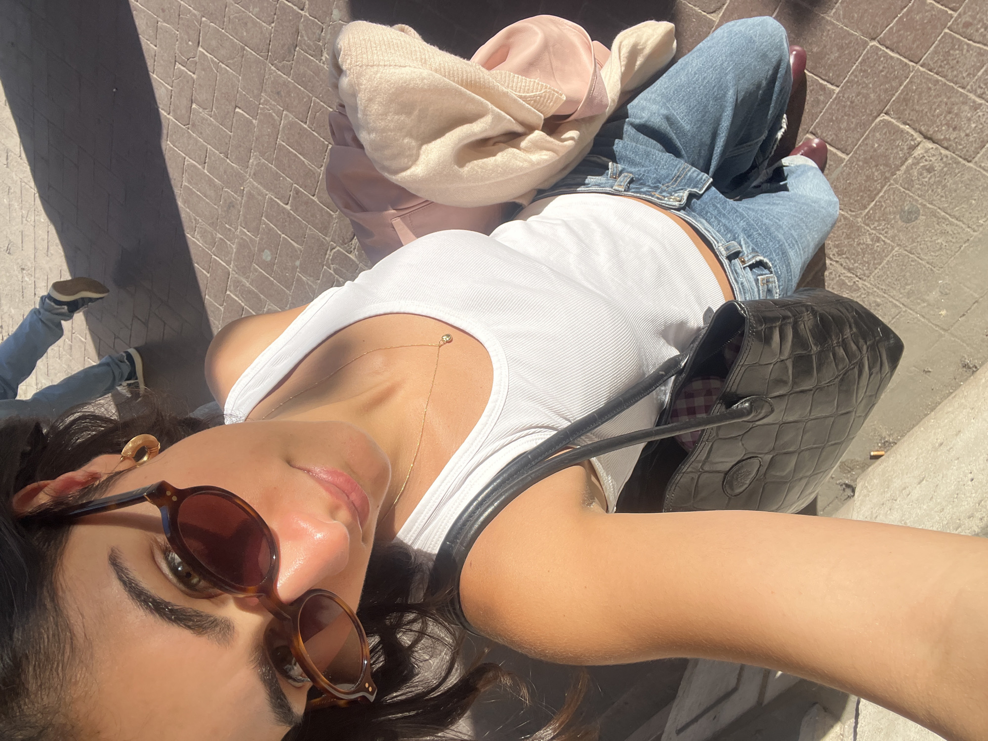 A woman is featured in a close-up, high-angle selfie, looking up at the camera. She is wearing a white ribbed tank top, light blue denim jeans, and dark red shoes. Her dark hair is visible around her face, and she has a golden chain necklace with a small charm, a golden ear cuff/earring, and brown, round-framed sunglasses on her face. Her lips are a soft pink.
She is holding a black, possibly quilted or embossed leather handbag, which has a distinct circular logo, likely Mulberry, visible on the flap. Draped over what appears to be her lap or a nearby surface are a light pink garment and a beige knitted sweater or scarf.
The setting is outdoors on a sunny day, indicated by strong shadows and bright highlights on her skin and the ground. The ground is paved with a herringbone brick pattern. In the background, to the lower left, part of another person's leg and foot are visible, wearing light blue jeans and light-colored sneakers. The scene conveys a relaxed outdoor moment, potentially in Paris, France, given the location context.