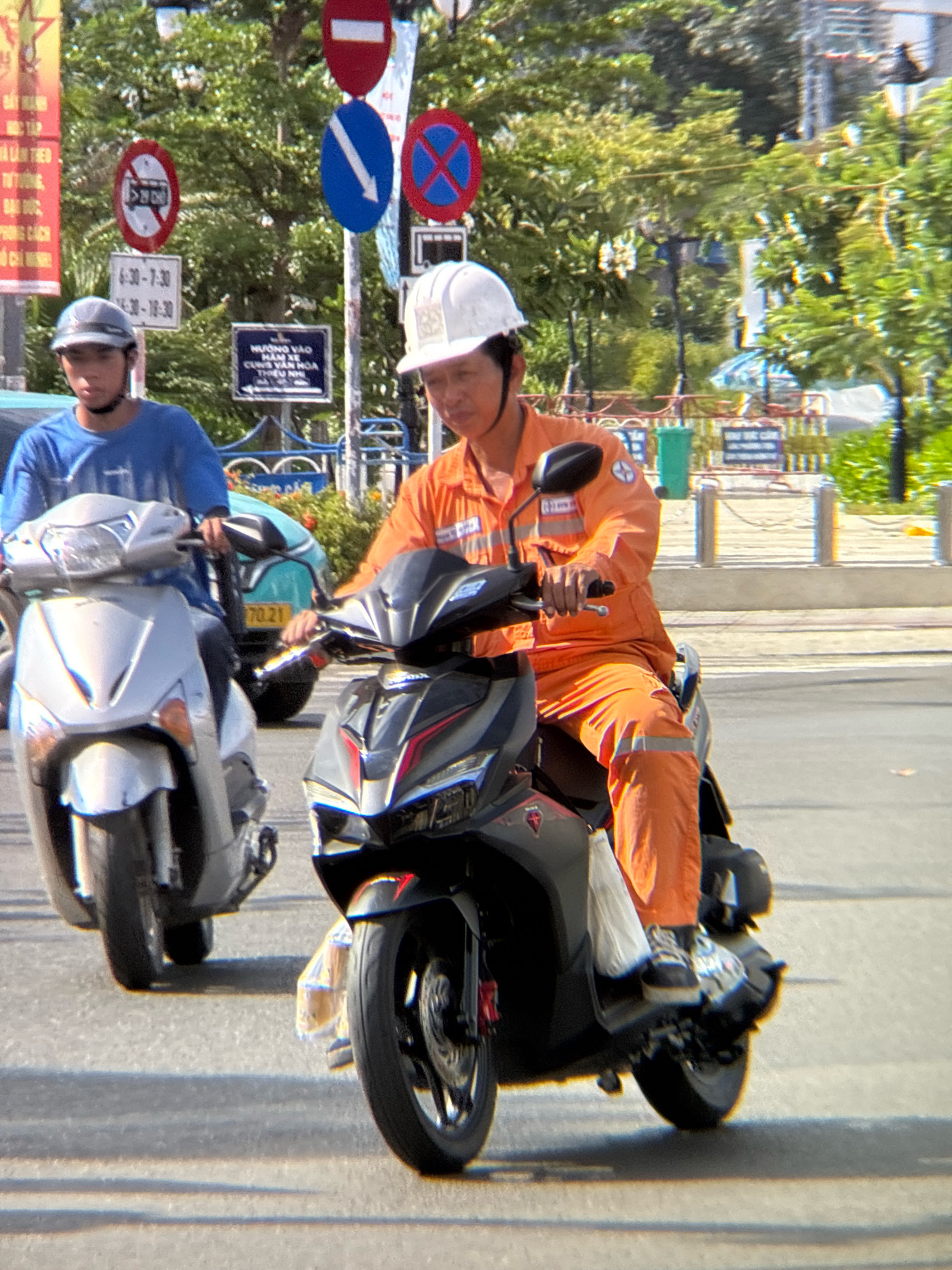 Two men are riding motorbikes on a paved road in Nha Trang, Vietnam. The man on the left, wearing a blue shirt and grey helmet, is riding a silver scooter. The man on the right, wearing an orange uniform and white helmet, is riding a black scooter. He is holding a white plastic bag. In the background, there are trees, street signs, and a green trash can. The street signs are in Vietnamese. One sign indicates "NO ENTRY 6:30 - 7:30" and "NO ENTRY 18:30 - 10:30". Another sign has a red cross in a blue circle, indicating "NO PARKING". The man on the right's orange uniform has reflective stripes on the lower legs. The lighting suggests it is daytime, with shadows cast on the road.