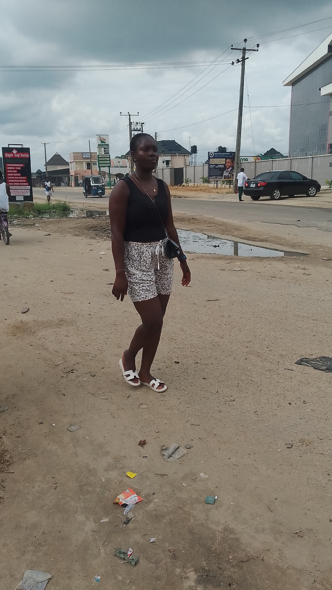 A young Black woman walks on a dusty, unpaved road in Warri, Nigeria, under an overcast sky. She is wearing a black tank top and patterned shorts, with white sandals. She has a dark crossbody bag and a thin necklace. In the background, there are buildings, a utility pole with wires, a black car, a tricycle, and several signs, one of which reads "Maple Leaf Suites." The ground is littered with debris, including trash, and there are puddles of water on the road.