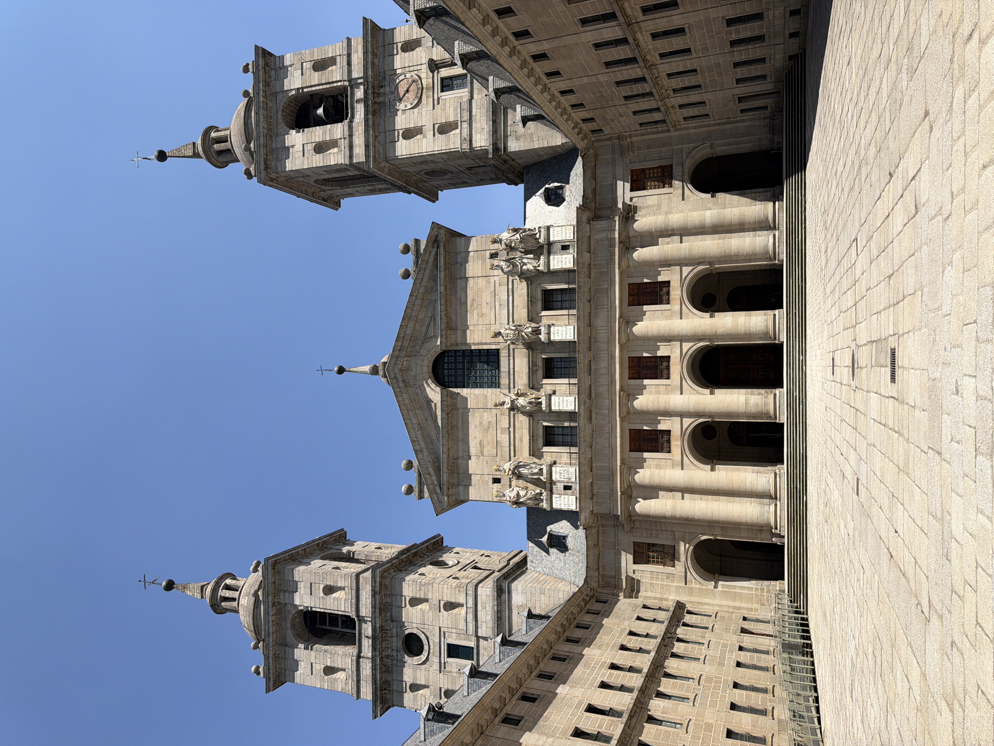 The image shows a symmetrical facade of a large, stone building, likely a royal monastery or palace, under a clear blue sky. Two identical bell towers flank a central, grander section of the building. The towers have arched openings, and each is topped with a pointed spire topped by a cross. The central section features classical architectural elements, including columns, arches, and statues of figures in regal attire adorning the upper level. The facade is constructed of light-colored stone blocks. In front of the building is a large, paved courtyard with intricate stone patterns. The courtyard is empty of people. The building complex extends outwards with additional wings, visible on the left and right sides of the frame, featuring rows of windows and dormer windows on the roof. The overall scene conveys a sense of grandeur and historical significance. This building is the Royal Site of San Lorenzo de El Escorial, located in San Lorenzo de El Escorial, Spain.