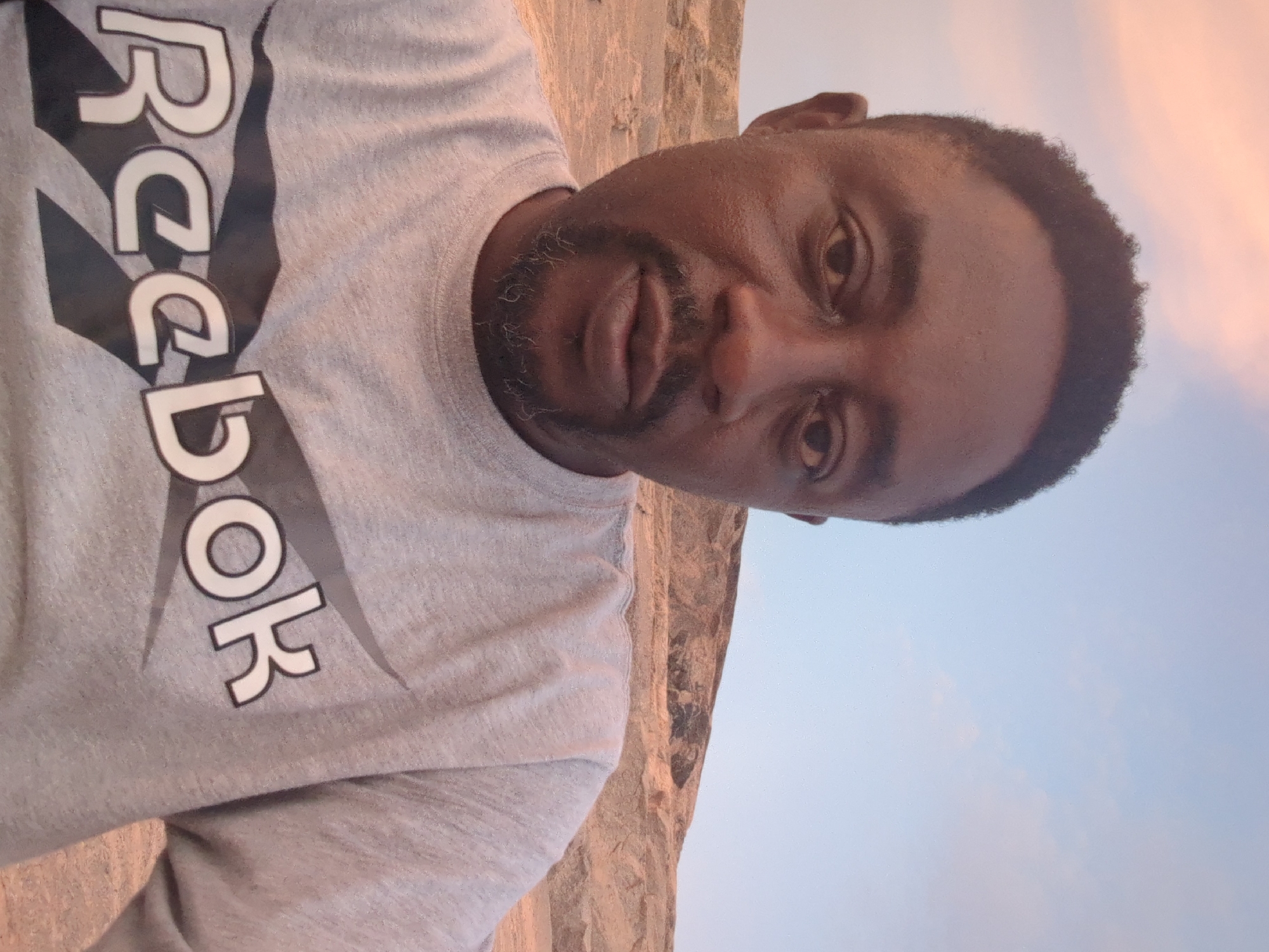 A medium-shot, portrait-style photograph shows a man wearing a gray Reebok-branded sweatshirt. The image is rotated counter-clockwise by approximately 90 degrees. The background suggests an outdoor setting with hints of a wall texture and a soft, gradient sky that transitions from a light blue to a faint pink, indicating either dusk or dawn. This image was taken in Narayi, Nigeria.