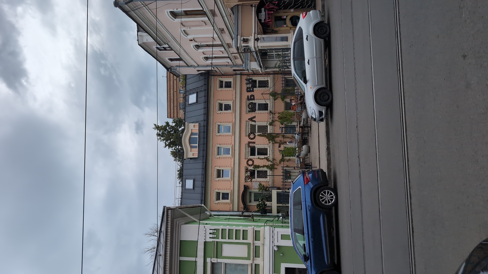 The image shows a street scene in Nizhny Novgorod, Russia. In the foreground, two cars are parked on the side of the road. A blue Skoda Kodiaq is parked on the left, and a silver Lada Granta is parked on the right. The cars are positioned on a paved road with trolley tracks running through it.
Behind the cars, a three-story building with a salmon-pink facade stands. The building has a dark grey mansard roof with several dormer windows. The facade is adorned with a sign made of illuminated letters spelling out "ЭТО ПО ЛЮБВИ" which translates to "This is out of love." The windows on the second and third floors are symmetrically placed and appear to be dark. The ground floor features an outdoor seating area with tables and chairs, suggesting a cafe or restaurant. Some patrons are seated at the tables. Green vines are draped across the facade.
To the left of the pink building is a narrower building with a vibrant green facade, decorated with white architectural details. To the right of the pink building is a lighter-colored building, possibly beige or light pink, with arched windows on the upper floors. The sky is overcast with dark clouds, indicating a cloudy day. The overall environment appears to be an urban setting with a mix of architectural styles.