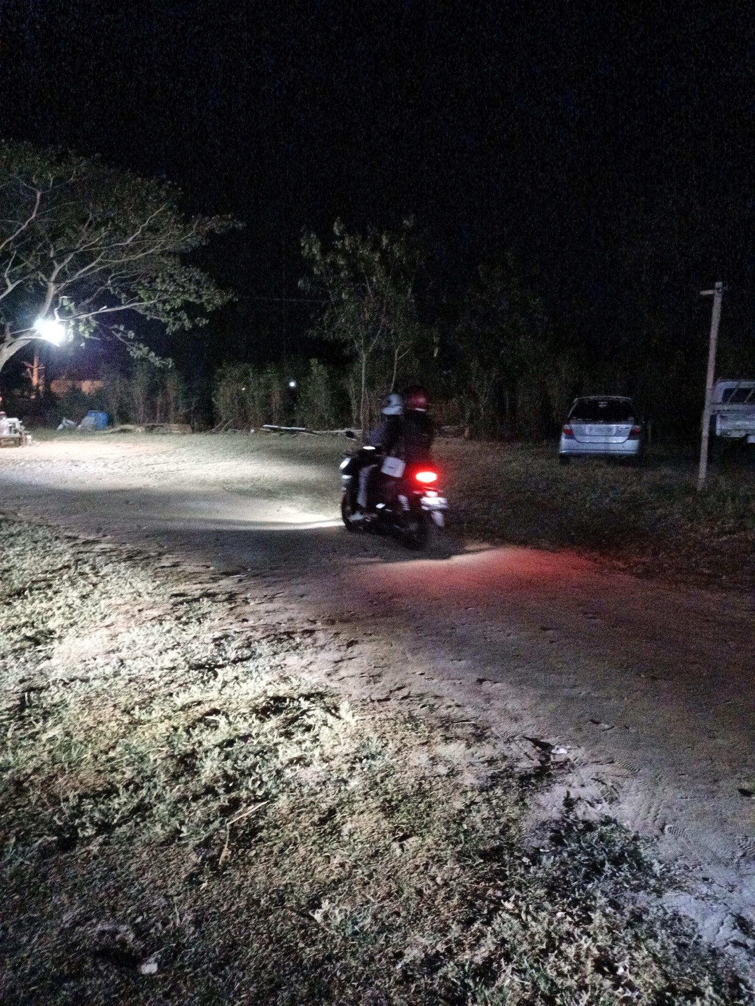 An exterior nighttime scene shows two people riding on a motorcycle on a dirt road. The motorcycle's taillight is illuminated red, and its headlights cast light on the road ahead. The motorcycle is moving from left to right in the frame. In the background, there is a silver car parked on the right side of the road, and to the left, there are trees and a tent. The ground is a mixture of dirt and grass. The scene appears to be in a rural area, possibly on the outskirts of Cikal, Indonesia, given the general ambiance.