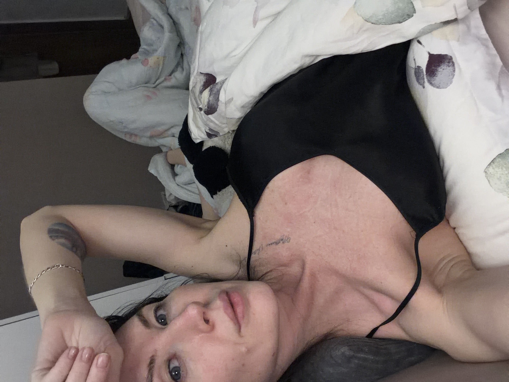 A fair-skinned woman with blue eyes and dark hair lies in bed. She is wearing a black top with thin straps. Her right arm is bent, with her hand partially covering her forehead. A silver bracelet adorns her right wrist. A tattoo is visible on her right forearm and a small tattoo is visible on her chest. She is partially covered by a white duvet with a watercolor-like design of plums. The bedding is rumpled. The location is likely a bedroom in a private residence. The city of Perm, Russia is not evident in the image.