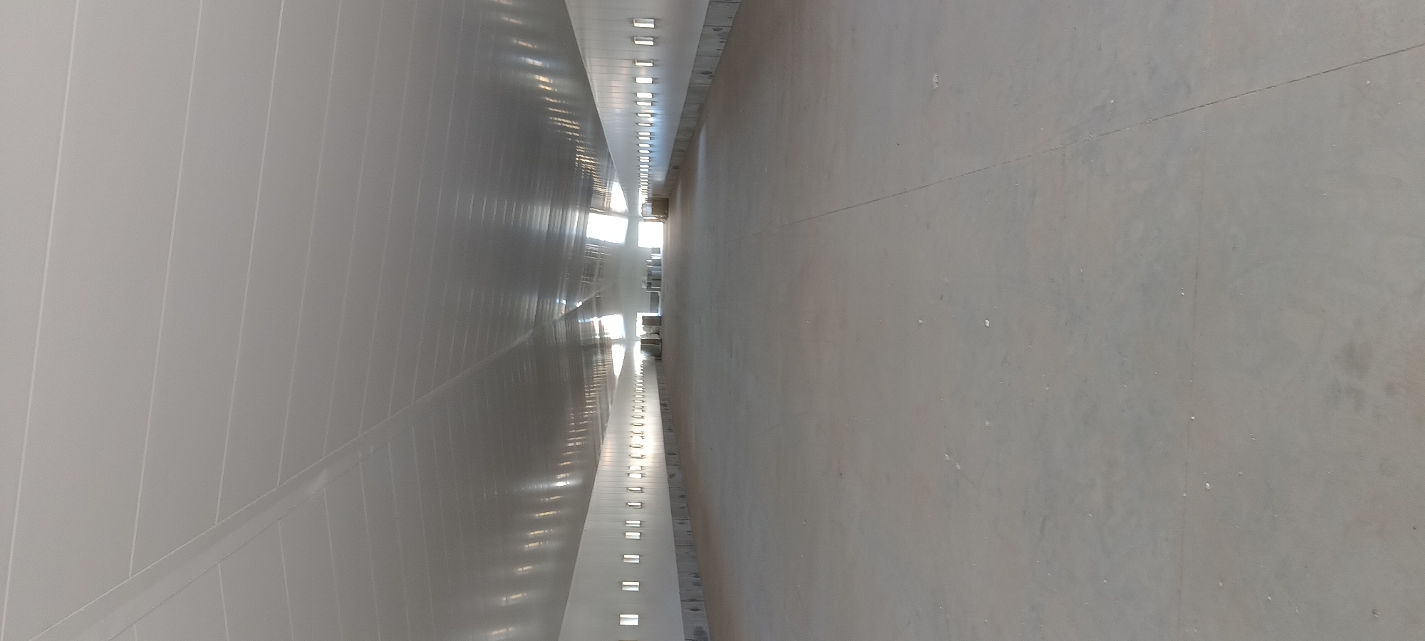 The image shows the interior of a large, empty, industrial building, likely a warehouse or agricultural structure, in Magnuszewice, Poland. The floor is a smooth, grey concrete surface. The walls are white, and along them are rows of small, rectangular windows. The ceiling is white and has a ribbed or corrugated texture, with visible beams or supports. Light streams in from openings at the far end of the building, illuminating the space and creating reflections on the ceiling. There are no people visible in the image. Some indistinct objects, possibly pallets or containers, are present in the distance. The overall impression is one of a new or recently completed construction.