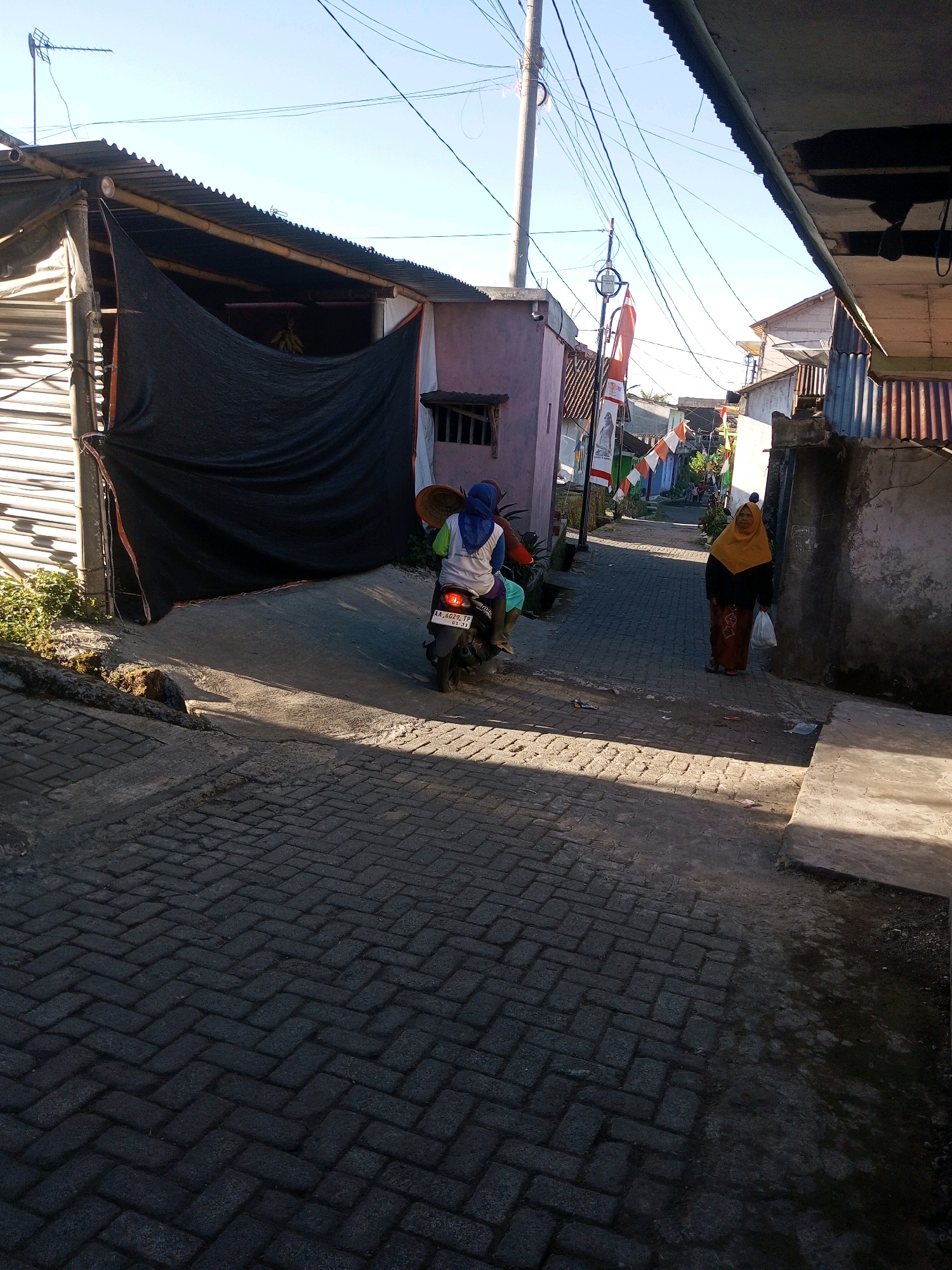 The image depicts a narrow street paved with interlocking bricks in Andongsili, Indonesia, bathed in morning sunlight. A person rides a motorcycle, wearing a blue headscarf and a green shirt, carrying a basket on the back of the bike. Another person, a woman wearing a yellow headscarf and a patterned skirt, stands on the right side of the street holding a white plastic bag. Buildings with corrugated metal roofs and varying facades line both sides of the street, some displaying Indonesian flags. Power lines crisscross the sky, and vegetation is visible on the left side of the image. The scene conveys a sense of everyday life in a rural or semi-urban Indonesian setting.