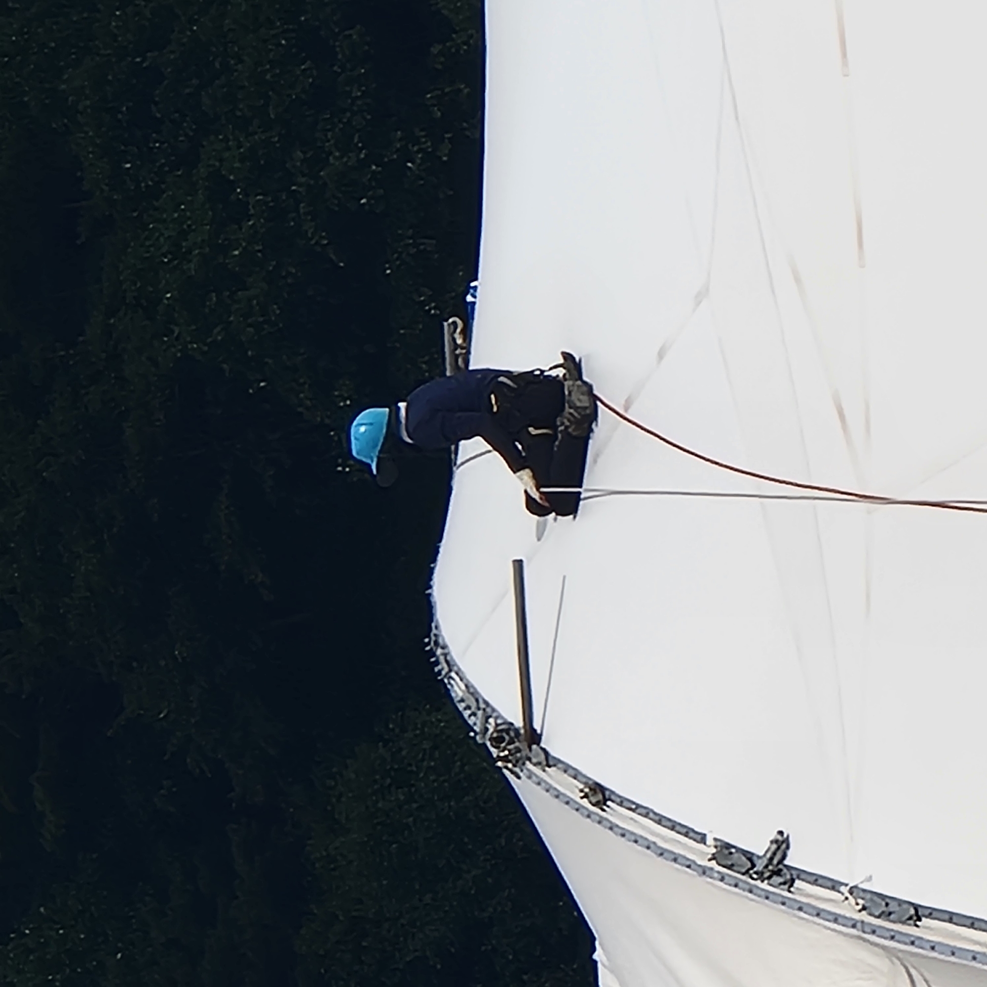 A person wearing a blue hard hat, dark blue long-sleeved shirt, dark pants, and work boots sits on a large white tent. The person is facing to the left and appears to be wearing a harness. A wooden pole is placed on the tent in front of the person. The tent is white with metal fasteners along its edge. In the background, dark green trees are visible. The scene appears to be outdoors.