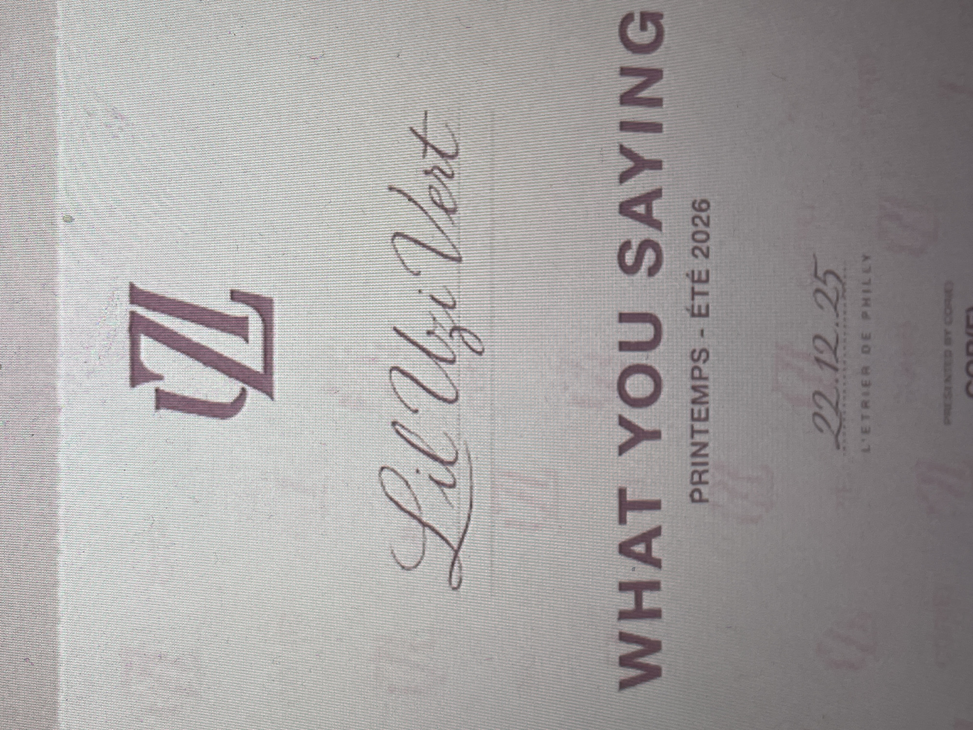 The image displays a photograph of what appears to be a promotional poster or screen, tilted counter-clockwise. The background is a light, textured off-white or beige, resembling a fine fabric weave or a digital screen texture.
The primary subjects are text and a logo. A prominent, stylized dark brown or maroon "Z" logo is positioned on the left side. To its right, the name "Lil Uzi Vert" is elegantly written in a flowing, dark cursive script. Below this, in large, bold, uppercase letters, is the phrase "WHAT YOU SAYING".
Further details include smaller text: "PRINTEMPS - ÉTÉ 2026" (Spring - Summer 2026) in a sans-serif font, suggesting a seasonal period and year. Below this, a date "22.12.25" is presented in a refined script font, likely referring to December 25, 2022. Underneath the date, "L'ETRIER DE PHILLY" (The Stirrup of Philly) is visible, strongly hinting at a connection to Philadelphia. In the bottom right corner, smaller text reads "PRESENTED BY CORIE" and "COPIED", indicating credits or copyright.
No people are visible in the image. The setting appears to be an indoor space where such promotional material would be displayed. The activity suggested is the announcement or promotion of a project or event related to the artist Lil Uzi Vert. While the photograph itself is located in Sofia, Bulgaria, the contextual information within the image points to Philadelphia. No specific time of day or weather can be discerned.