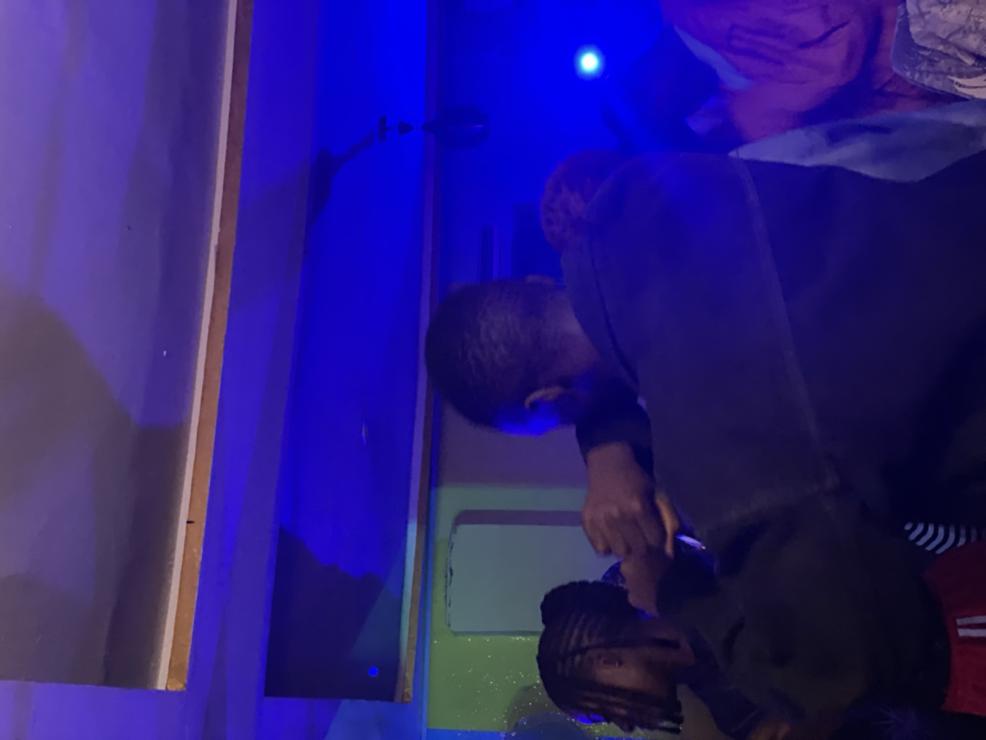 The image captures an indoor scene, likely a gathering or event, illuminated by vibrant blue and green lighting. In the foreground, a person is seen from behind, wearing a dark jacket. To the left, another individual with braided hair is partially visible, facing away from the camera. The background features walls and a ceiling with architectural elements, suggesting an interior space. The lighting creates a dynamic and atmospheric effect, with shadows and reflections playing across the surfaces. The overall impression is one of a lively, possibly nocturnal, social setting. There is no explicit information in the image to determine its location within Durumi, Nigeria.