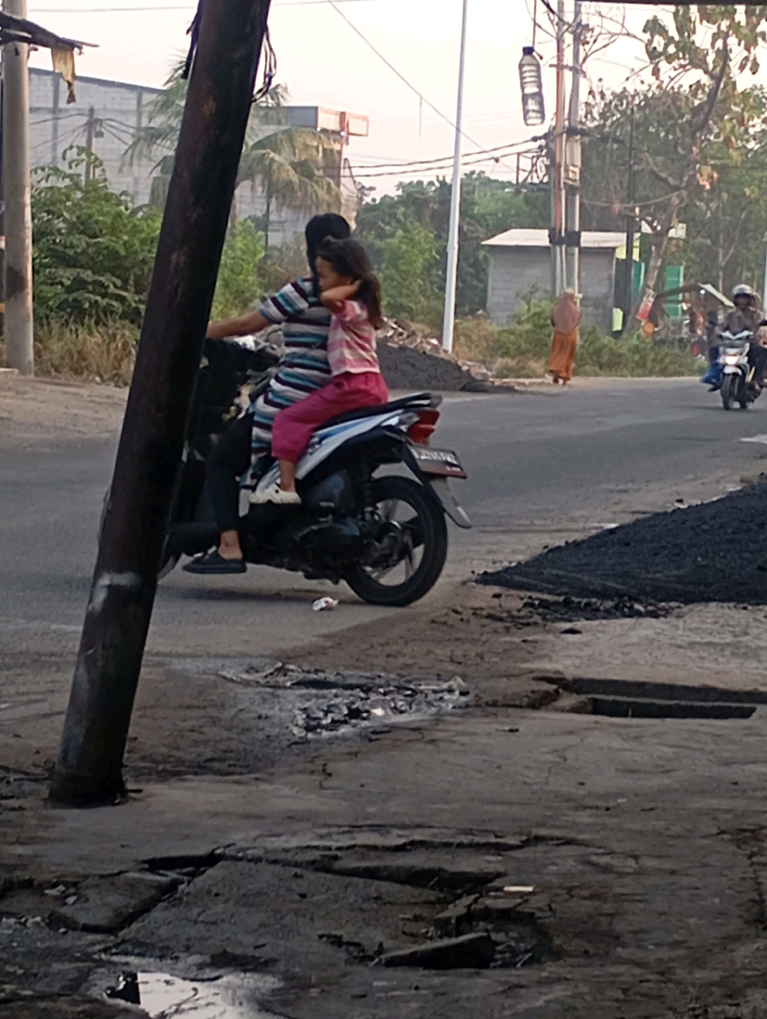 The image depicts a street scene in Babelan, Indonesia, with a focus on a motorcycle carrying two individuals. A woman in a striped shirt is driving the motorcycle, with a young girl sitting in front of her. The girl is wearing a pink striped shirt and pink pants and has her arm around the woman's neck. They are traveling on a paved road. In the background, there are several buildings, some with signage, and lush green trees. A person in an orange garment is walking away from the viewer in the mid-ground. Another motorcycle carrying two people is visible further down the road. Piles of dark, coarse material, possibly asphalt or gravel, are present on the side of the road, suggesting ongoing construction or road work. The overall lighting suggests it is daytime, likely late afternoon due to the soft light. The foreground shows a cracked and weathered asphalt surface, indicating the road's condition. A utility pole stands prominently in the left foreground, partially obscuring the view of the motorcycle.