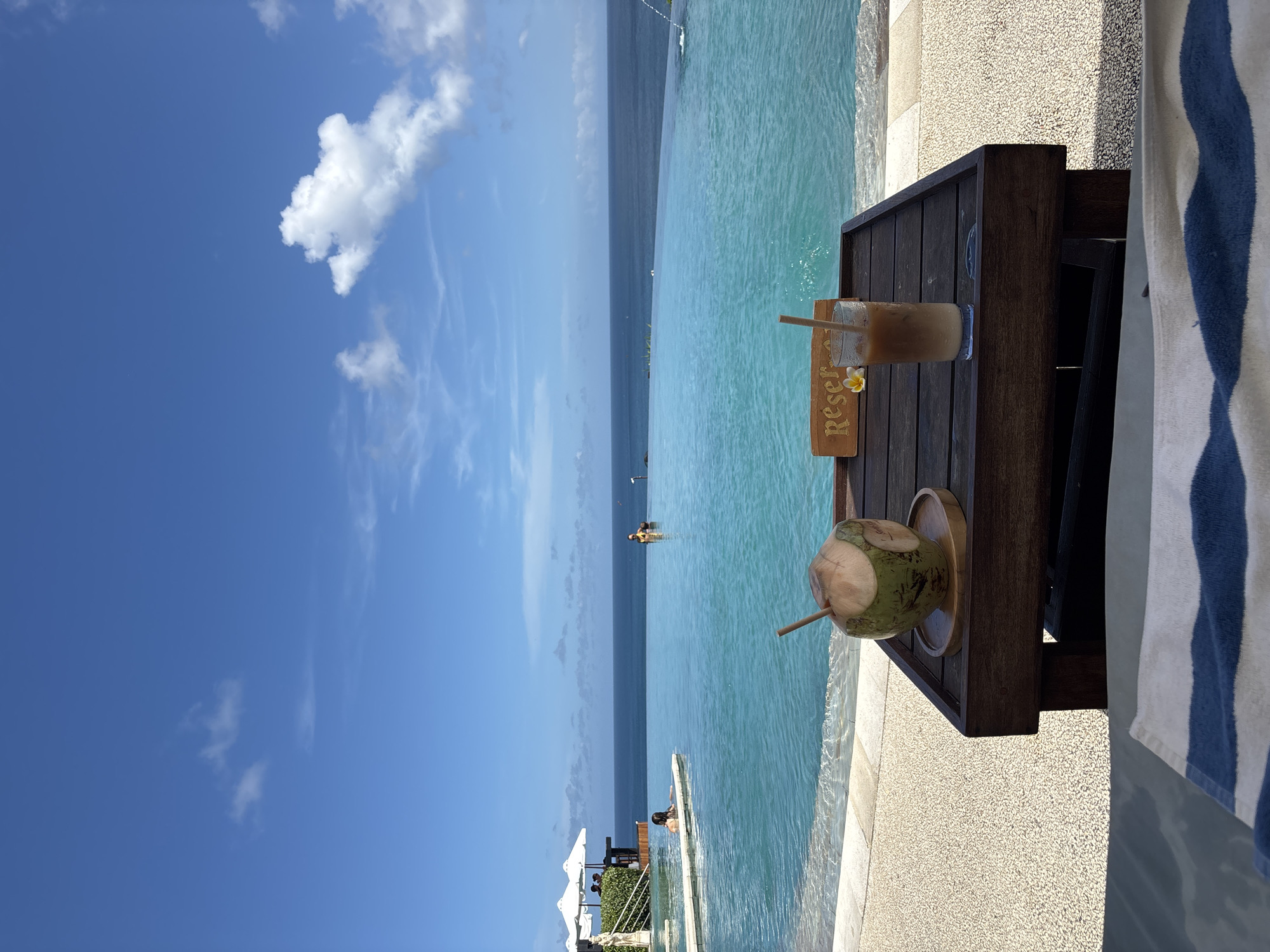 The image shows a view overlooking an infinity pool merging with the ocean, under a blue sky with scattered clouds. A wooden table is positioned beside the pool, holding a coconut drink and a glass of iced coffee with a "Reserved" sign and a frangipani flower. In the background, people are visible swimming in the pool and standing near white umbrellas. The scene appears to be taken at a resort or hotel in Kuta Selatan, Indonesia, known for its beach and ocean views. A striped towel lies next to the table.