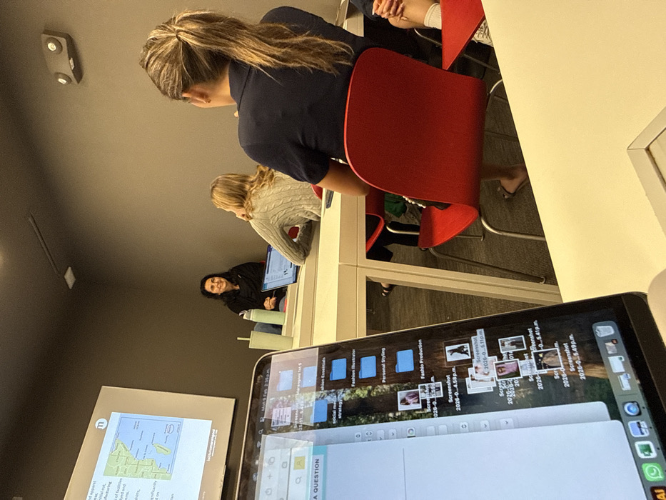 A classroom scene shows three students at a table. The first student, with blonde hair pulled back, wears a navy shirt and sits in a red chair. The second has a gray sweater. The third, in a black hoodie, is smiling. A laptop screen displays files like "Fashion Illustration" and "Fashion Production". Another screen on the left shows a map and text, with the bottom corner reading "A QUESTION".
