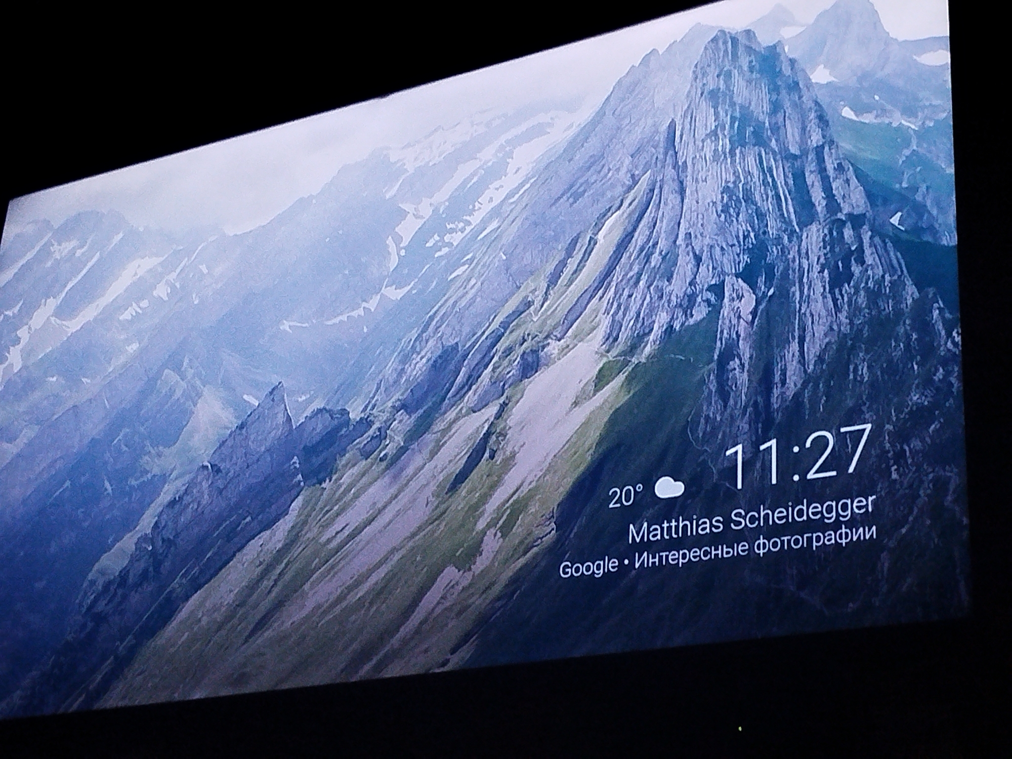 The image displays a large, high-definition screen showing a mountainous landscape. The screen is angled, with a black border visible on the top and sides. On the screen, there is text overlayed on the landscape. The text includes a time of "11:27", a temperature of "20°" with a cloud icon, and the name "Matthias Scheidegger". Below this, there is text in Russian: "Google • Интересные фотографии" which translates to "Google • Interesting photos". The landscape itself is characterized by steep, rocky mountains with patches of snow and green vegetation. The lighting suggests it is daytime, with a hazy or overcast sky. There are no people or discernible objects that would place this image in Nazran, Russia.