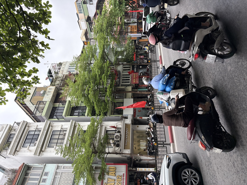 This image depicts a bustling street scene in Hà Nội, Vietnam, under an overcast sky. The foreground features three active motorbikes and a partial view of a white SUV on a damp asphalt road.
From left to right in the immediate foreground:
1. A white SUV's front left side and wheel are visible, positioned at the far left edge of the frame.
2. A dark-colored scooter, possibly a Vespa-style, is ridden by an individual wearing a dark brown long-sleeved shirt, dark pants, and a dark helmet. The scooter displays a rear license plate "29P1-523.44."
3. A black motorbike, positioned centrally, is operated by a person wearing a light blue long-sleeved shirt and a light-colored helmet. This motorbike is heavily loaded with multiple large, blue cylindrical bundles and a white sack secured to its rear rack.
4. A silver-white scooter is ridden by an individual wearing a dark jacket, dark pants, a pink helmet, and a black backpack. Its rear license plate reads "90-B4 049.96."
The midground features a black metal barrier separating the main traffic lanes from an elevated sidewalk or parking area. Numerous additional motorbikes are parked behind this barrier, and several more are in motion further down the street, including one rider wearing a "Grab" branded jacket.
The background is dominated by multi-story, narrow buildings characteristic of urban Vietnamese architecture. These structures exhibit varying states of maintenance and include commercial establishments on their ground floors. Visible storefronts and signs feature Vietnamese text, such as "HỌC VIỆN TÓC CHÂU ÂU - 292," "THU KENNY MAKE-UP," and "DỊCH THUẬT MINH QUÂN." Several red flags, consistent with the national flag of Vietnam or the Communist Party flag, are mounted on balconies or poles extending from the buildings. Green, leafy trees are planted along the street, their foliage partially obscuring some building facades and providing an overhead canopy in the upper right quadrant of the image. The sky is a uniform, bright grey, indicating diffused daylight without direct sunlight.