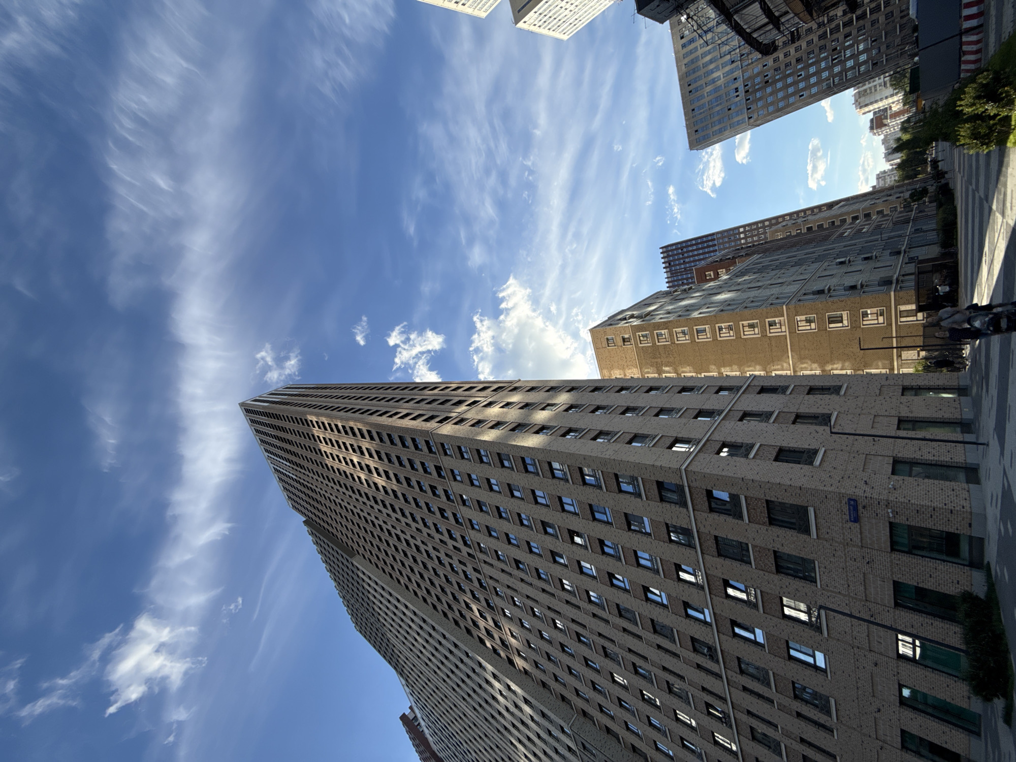 This is an image taken from a low angle, looking up at a cluster of modern high-rise apartment buildings in Moscow, Russia. The sky is a vibrant blue, dotted with wispy cirrus clouds. The buildings are tall and angular, with a mix of beige brick and darker facade materials. Many windows are visible, reflecting the light.
In the foreground, there is a paved walkway with a checkered pattern. Several people are walking on the path, some with strollers or bicycles. There are also small green bushes and trees lining the walkway and in front of some of the buildings. The overall impression is of a contemporary urban residential area. The building on the far left of the image is the tallest and most prominent, with a light beige brick facade and numerous rectangular windows. To its right are other buildings of varying heights and architectural styles, creating a dynamic cityscape. The lighting suggests it is daytime, with sunlight casting shadows.