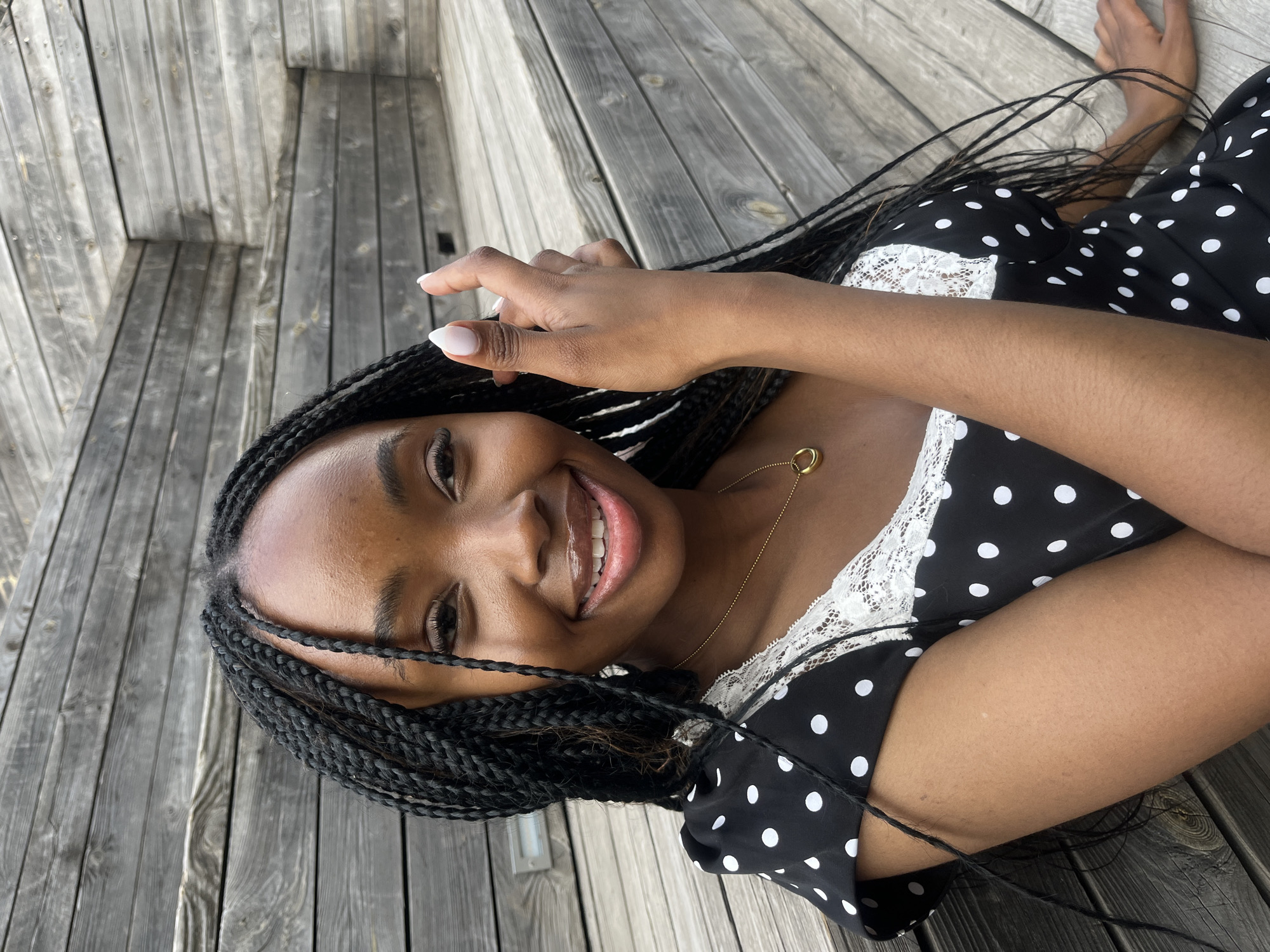 A young Black woman with braided hair is seated on wooden steps outdoors. She is wearing a black dress with white polka dots and white lace trim around the neckline. She has a gold necklace with a circular pendant. Her left hand is raised near her face, and her right hand is resting on the steps. The background consists of weathered wooden planks arranged as steps and walls. There is no indication of the city of Kazan, Russia, in the image.
