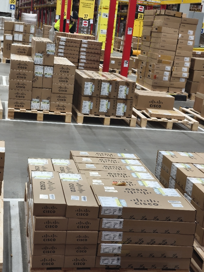 The image depicts the interior of a large industrial warehouse or distribution center, likely situated in Beringe, Netherlands. The floor is a light grey concrete marked with white painted lines. Numerous brown cardboard boxes, prominently displaying the "CISCO." logo and text, are stacked on standard wooden shipping pallets across the foreground and midground. Many boxes feature white labels with black text and barcodes. In the background, tall metal industrial shelving units, characterized by red vertical support beams, are loaded with additional boxes. Yellow vertical banners or signs, some with "ATTENTION" text and numerical designations such as '35', '37', and '38', are affixed to the shelving. A red circular 'no entry' sign is visible below the '37' marker. One individual is present in the midground, wearing a high-visibility yellow vest over dark attire, facing away from the camera and appearing to be engaged in activity near the shelving units. The environment is brightly lit and organized, indicative of a logistics operation.