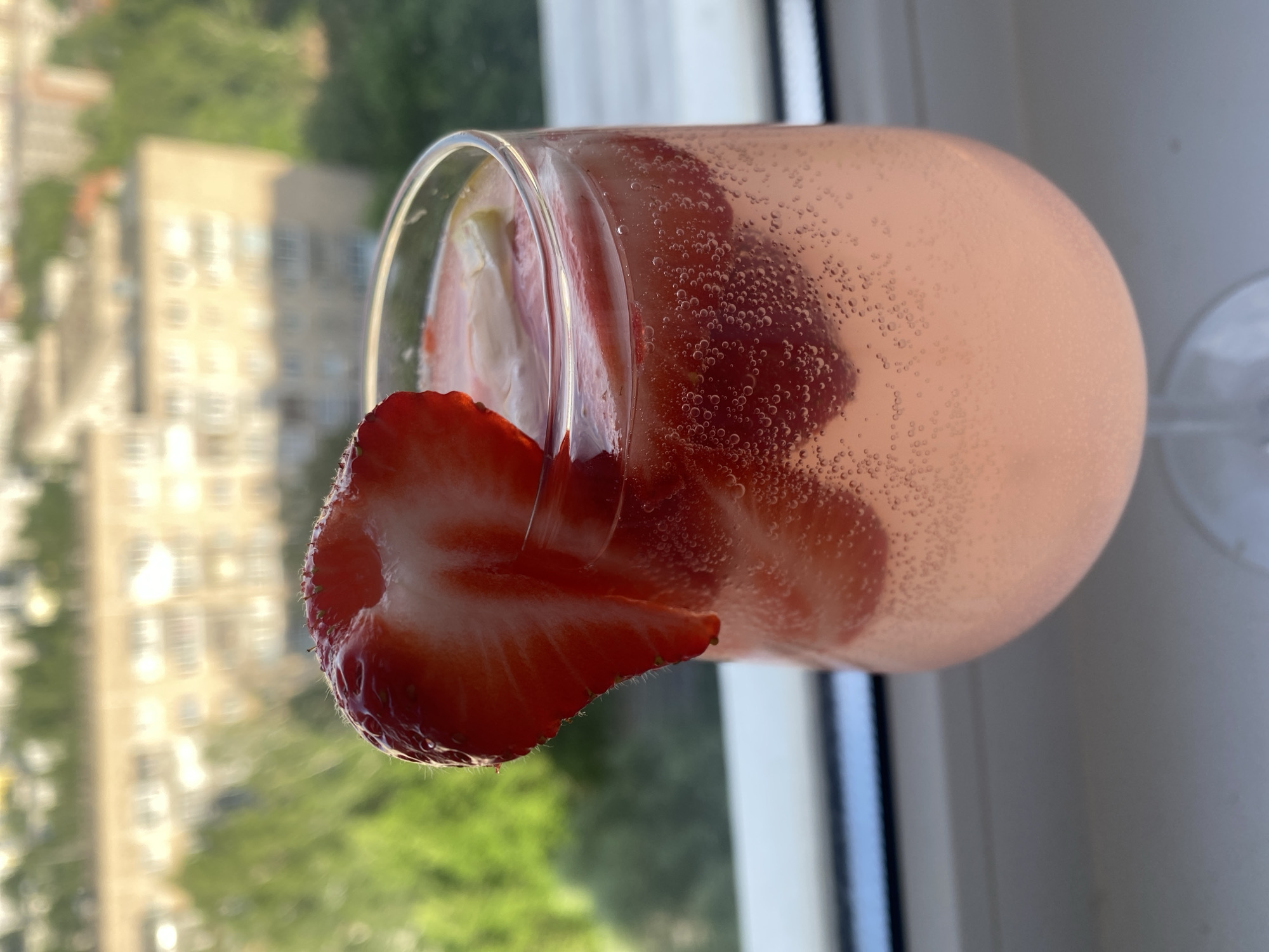 A close-up, eye-level shot shows a glass of pink cocktail garnished with a slice of strawberry. The glass is filled with a bubbly beverage, and a thick slice of fresh strawberry is positioned on the rim, with the cut side visible. The background features a blurred cityscape, seemingly viewed through a window. The setting is a residential building located in Izhevsk, Russia. The light suggests daytime. The environment includes the window frame. There are no people present in the frame.