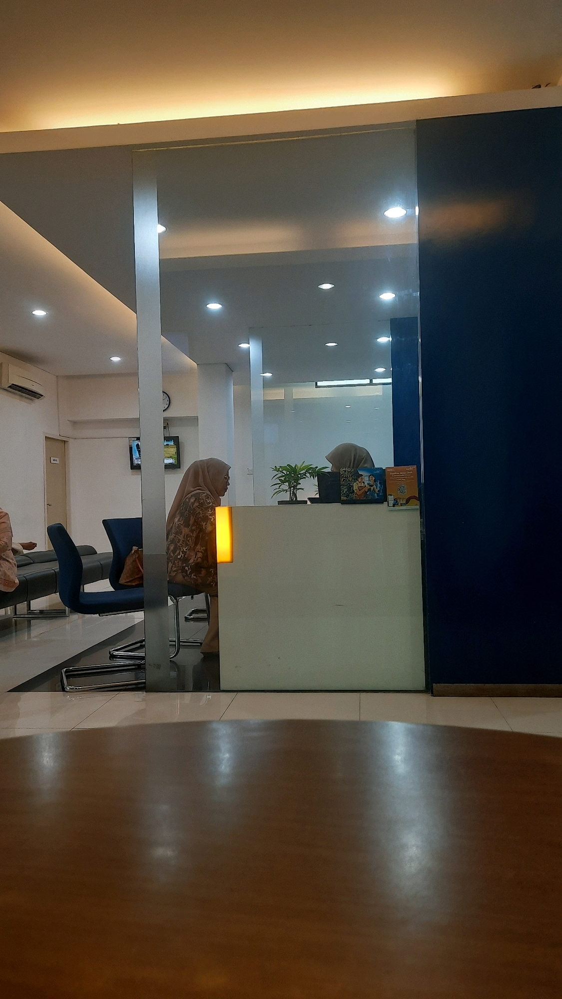 The image depicts the interior of what appears to be a waiting area or reception. In the foreground, a dark wooden surface, possibly a table, is visible. The background shows a reception desk with a person wearing a hijab seated behind it. To the left of the desk, another person wearing a hijab is seated on a chair, facing away from the camera. Several chairs and a plant are also visible. The environment appears to be a modern office or service establishment, with overhead lighting and reflective surfaces. The overall lighting is soft and warm. The image does not contain any discernible elements that would specifically place it in Padang, Indonesia.