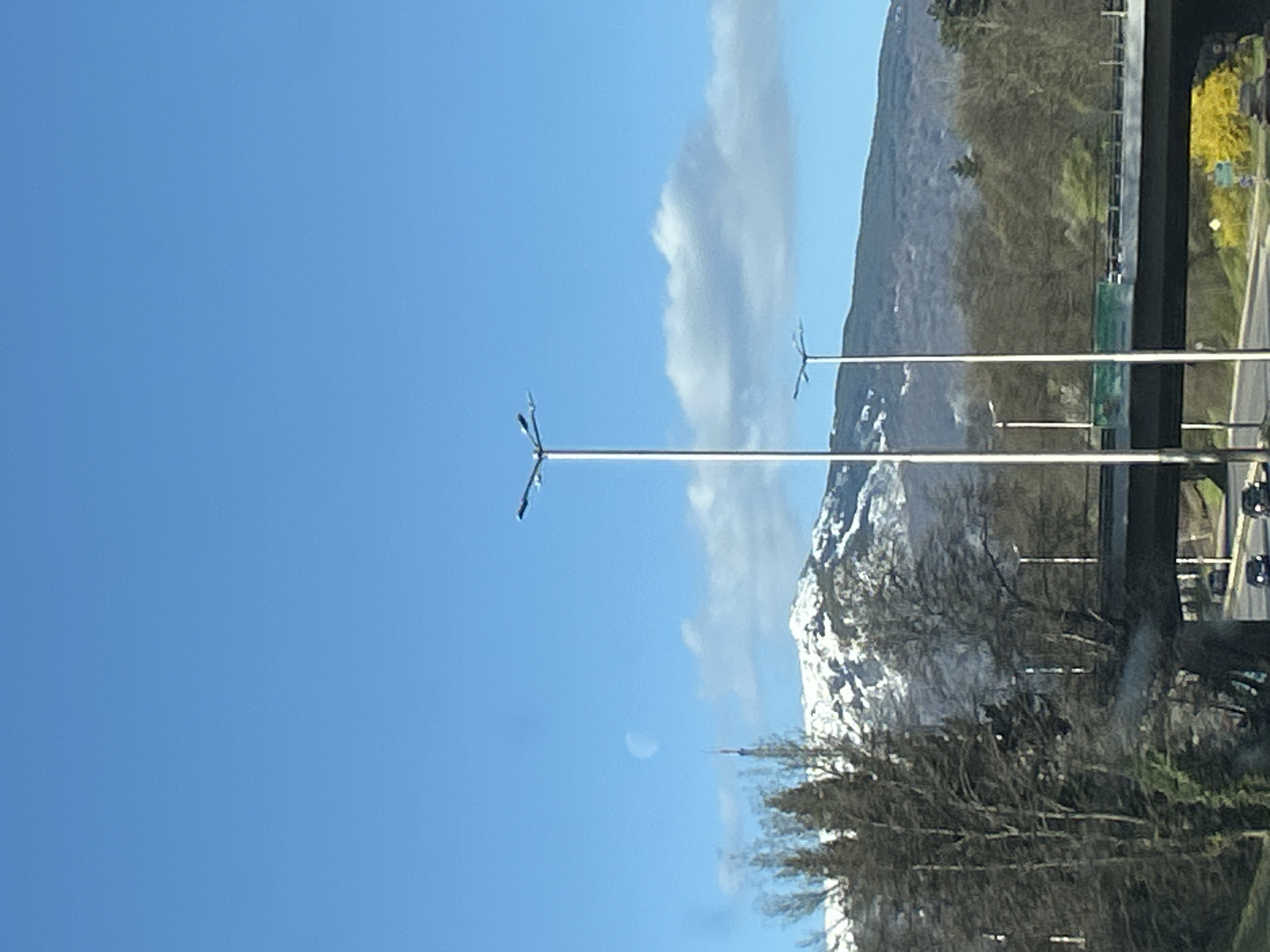 Here is a description of the image:
The image shows a view of a mountain landscape with a clear blue sky. In the foreground, there are two tall light poles, and beyond them, a snow-capped mountain. There are trees and vegetation at the base of the mountain. Additionally, an overpass with cars is visible to the right of the mountain. The location appears to be a highway or road. The image is taken from a moving vehicle.