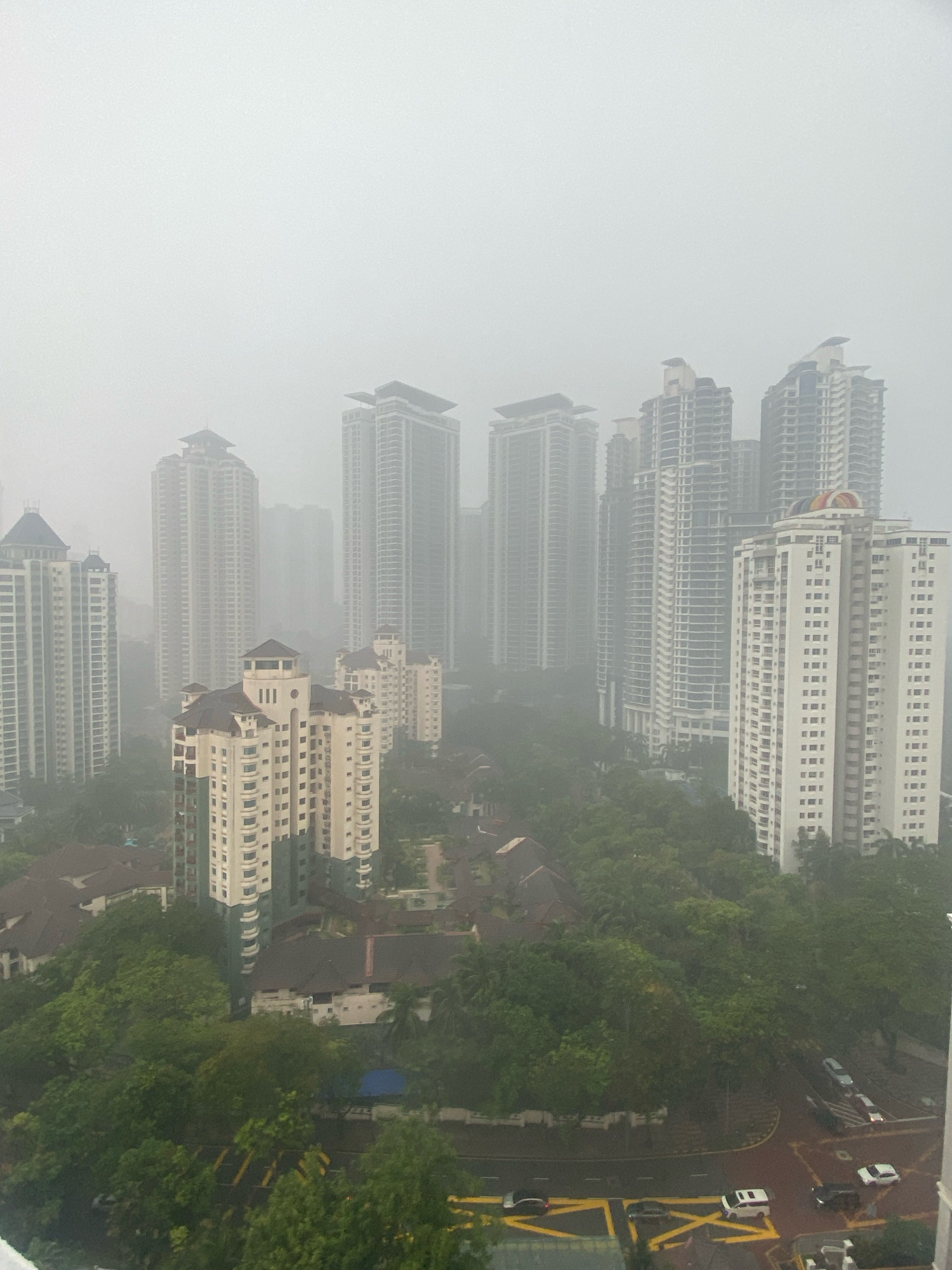 The image depicts a dense urban environment in Kuala Lumpur, Malaysia, shrouded in heavy mist or fog, significantly reducing visibility. A cluster of high-rise apartment buildings and condominiums dominates the skyline. The architecture of these buildings is characterized by modern, sleek designs with numerous balconies and repetitive window patterns. Some of the buildings appear to be under construction or are unfinished, with exposed structural elements. Lush green trees and vegetation are interspersed among the buildings, suggesting a well-developed residential area with considerable landscaping. The lower portion of the image shows roads with a few vehicles, including cars and a van, navigating the misty conditions. The overall atmosphere is one of urban density with a prevalent atmospheric haze, which is common in Kuala Lumpur due to its tropical climate and occasional industrial pollution or haze events.