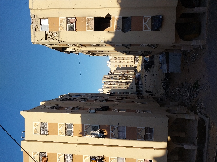 This image captures a damaged urban scene in Khan Yunis, Palestinian Territory, looking down an alleyway between two multi-story apartment buildings under a clear blue sky. The left building, light beige with decorative white window grilles, displays clothes hanging from several windows and balconies, suggesting continued occupancy. The right building, architecturally similar, exhibits extensive structural damage on its upper floors, including large missing wall sections, exposed interiors, shattered windows, and numerous visible impact marks, consistent with bullet or shrapnel damage, on its facade.
The unpaved ground between these buildings is covered with considerable rubble, dirt, and debris. Several dark figures, including adults and smaller individuals consistent with children, are present in this debris-laden area, moving or standing. A makeshift shelter constructed from white tarpaulin and another dark, undefined structure are situated amidst the rubble on the right side of the ground level. In the background, a cluster of similar multi-story apartment blocks, also appearing damaged, is visible. Thin utility wires crisscross the upper sky, and long building shadows dominate the foreground ground level, particularly on the right side.