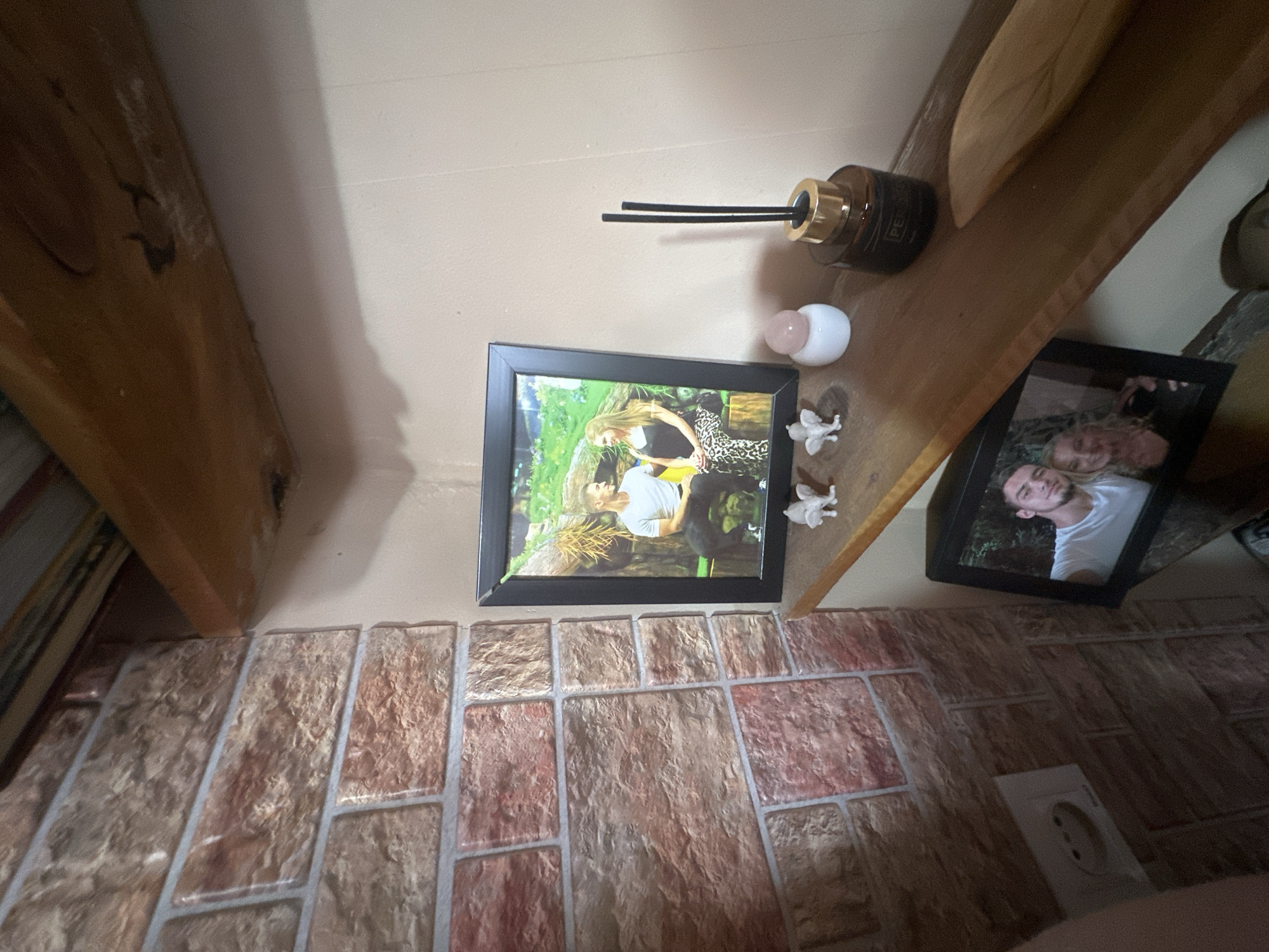 An indoor, medium shot captures a corner of a room featuring a brick-patterned wall and a wooden shelf. On the shelf, two framed photographs are displayed. The upper frame contains a picture of a man and a woman. To the left of this frame, there are two small white figurines, possibly angels, and a small white object topped with a pink sphere. To the right of the upper frame sits a dark glass bottle with a gold lid and black reeds, a reed diffuser. Below this shelf, a second framed photograph shows a man and a woman. The brick-patterned wall on the left has a white electrical outlet. The environment appears to be a domestic setting. The location is not specified within the image.