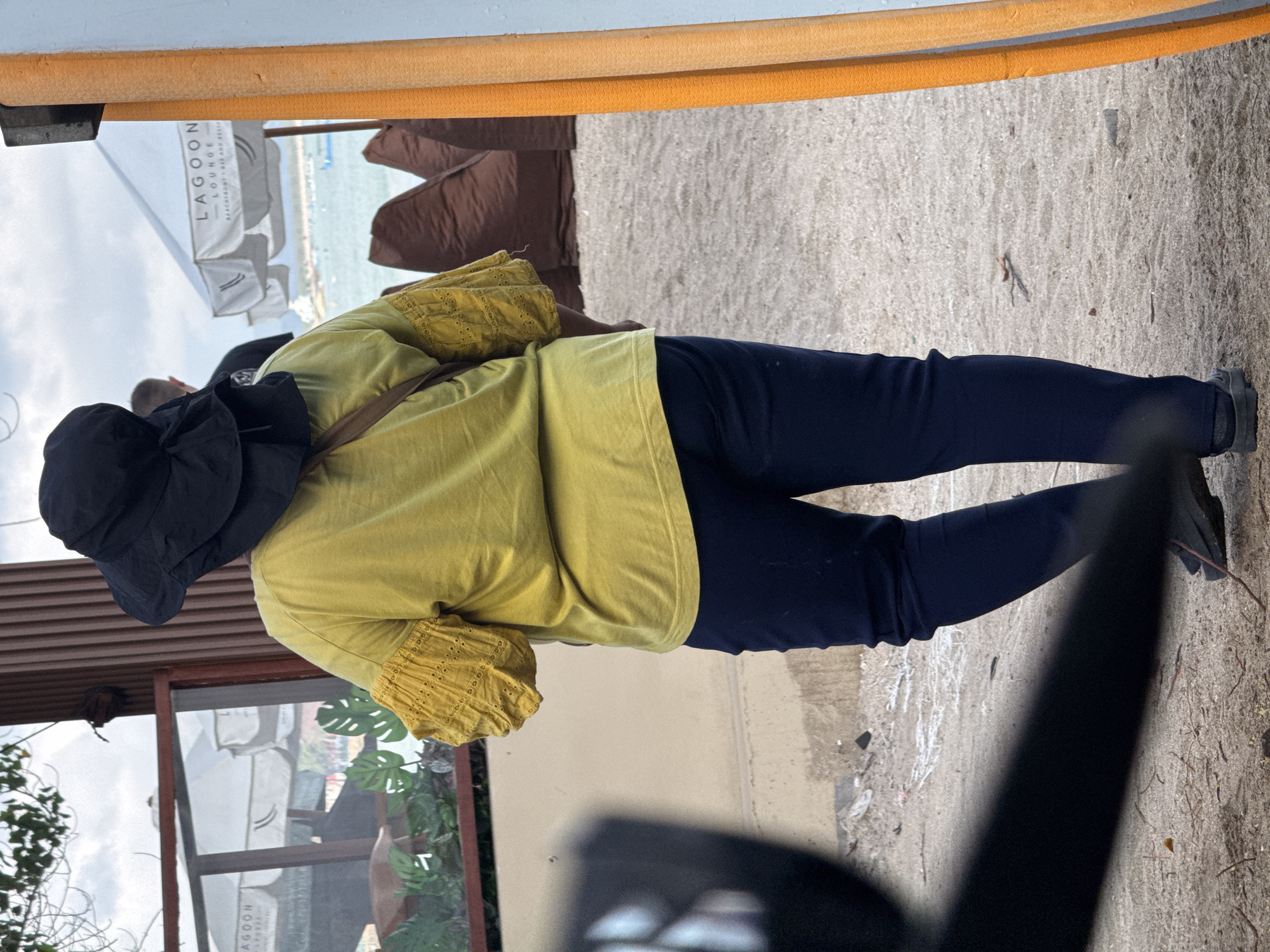 A person wearing a black sun hat, a yellow shirt with detailed sleeves, and dark blue pants walks away from the camera on a sandy surface. The person has a brown strap across their back. In the background, there is a beach scene with umbrellas and beanbag chairs, and water with boats. To the right, there are two orange textured posts. To the left, there is a dark, out-of-focus object. The location is Kuta, Indonesia.