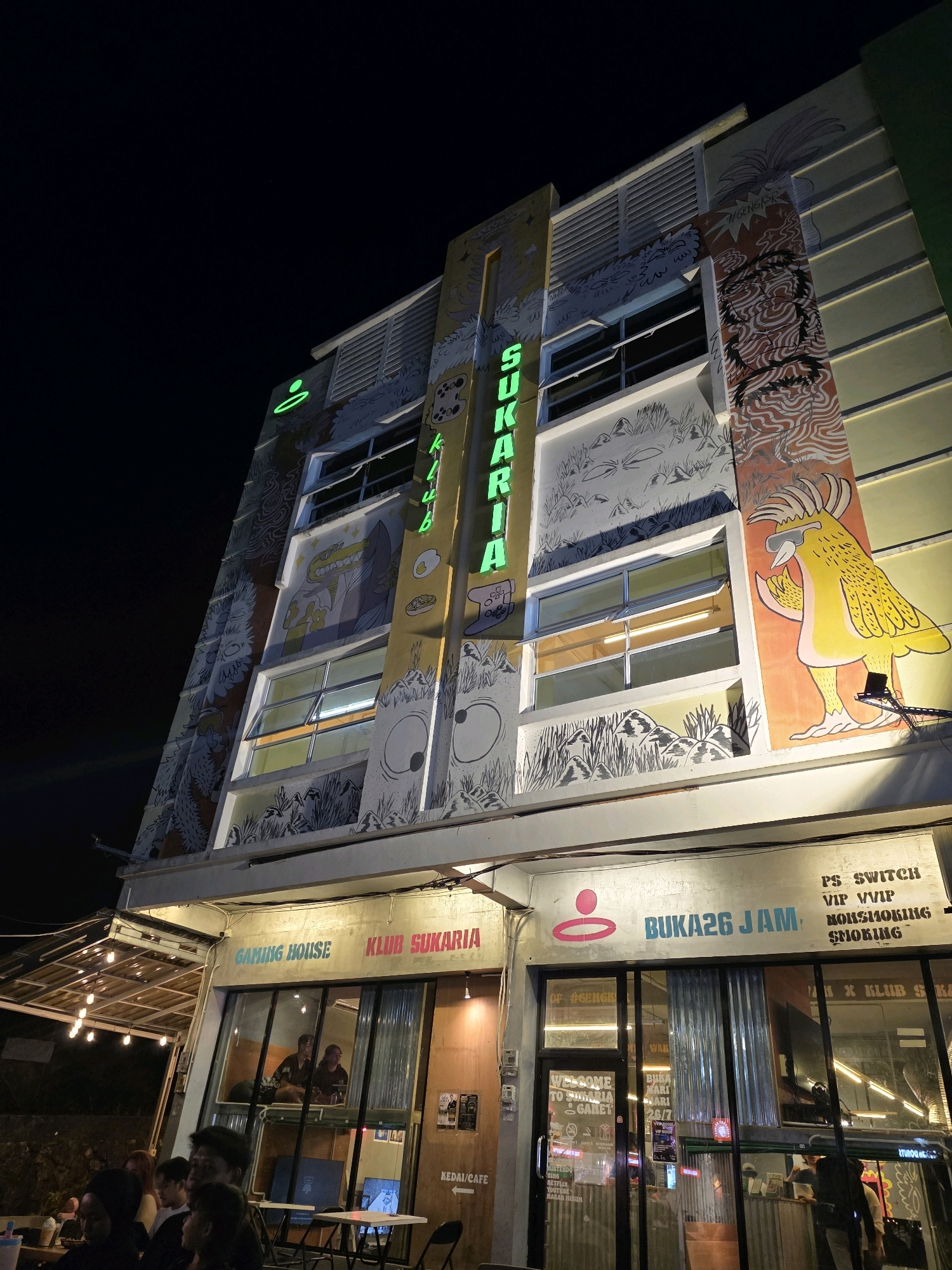 The image shows a multi-story building at night, likely in Tanjung Pinang, Indonesia, as indicated by signs. The building features a vibrant mural covering its facade, depicting cartoonish characters and natural elements. Neon signs, one in green spelling out "SUKARIA" vertically, are illuminated. The lower level of the building appears to be a commercial establishment with large glass windows and signage indicating "GAMING HOUSE KLUB SUKARIA" and "BUKA26 JAM" (Open 24 Hours). Inside the establishment, people can be seen through the windows. In front of the building, there are outdoor seating areas with string lights and people gathered. The overall scene suggests a lively evening at a recreational or entertainment venue.