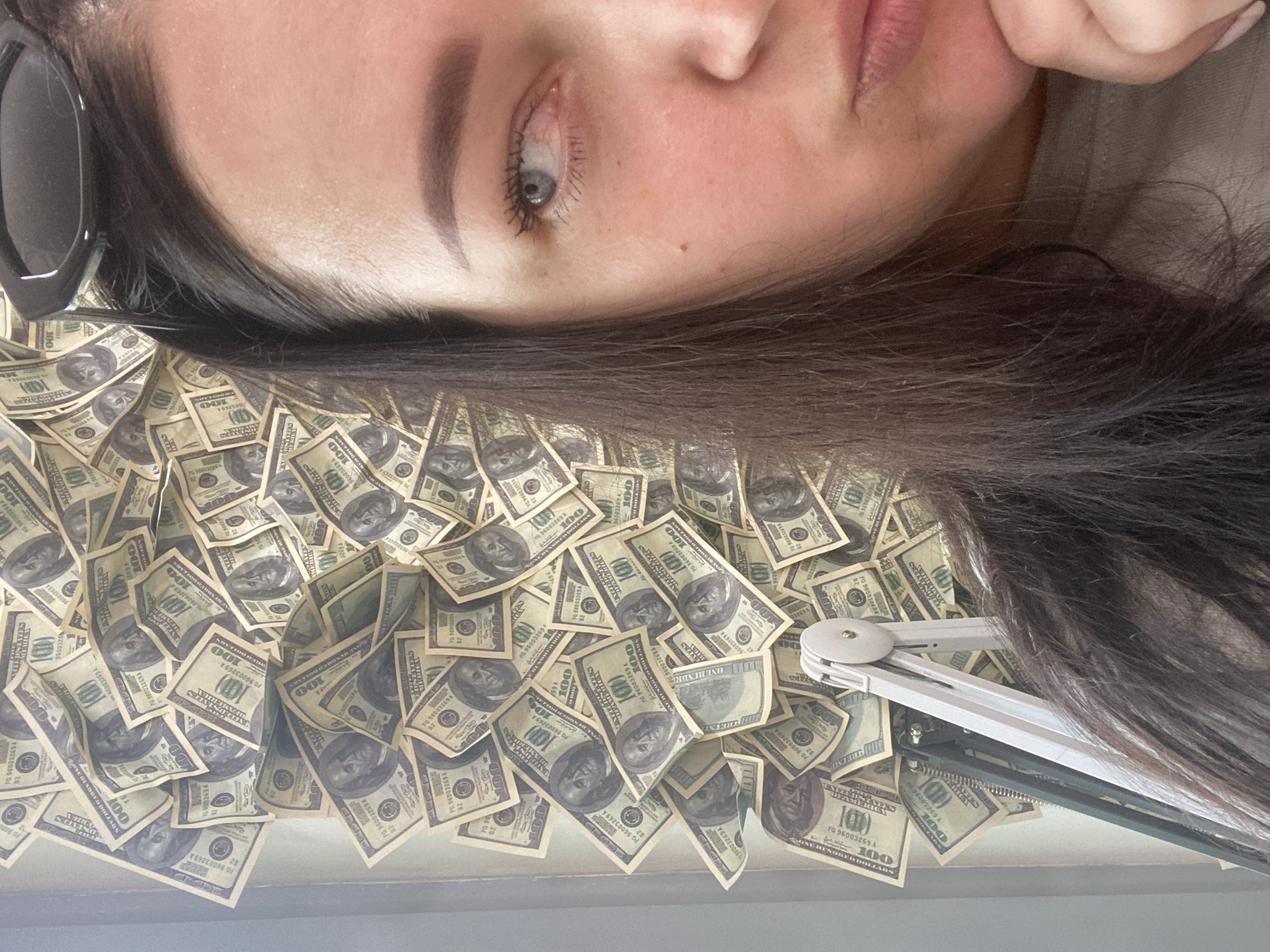 A close-up, upside-down, eye-level shot depicts a woman with dark hair lying on a surface covered with numerous one hundred dollar bills. She has fair skin, gray eyes, and neatly groomed eyebrows. She is also wearing sunglasses. Her right hand is positioned near her face, and she appears to be resting her head. An adjustable arm lamp is also visible.
The location and city of Sochi, Russia, cannot be verified from this image alone.