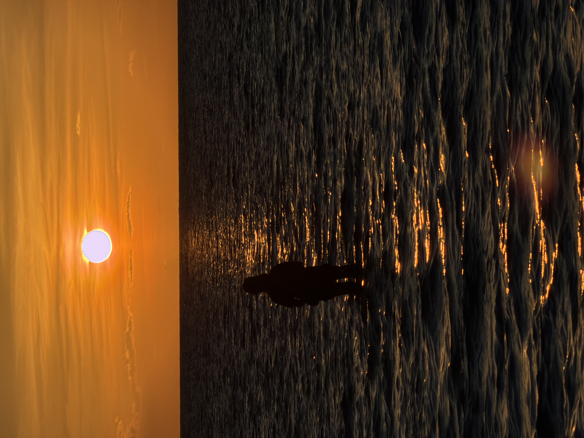 A single person stands in the water at sunset in Baan Plai Laem, Thailand. The sun, a bright white orb with a pale purple halo, is positioned in the upper center of the image, casting a warm orange glow across the sky. The horizon line is sharp and clear, separating the vibrant sky from the dark, rippling water. The water reflects the sun's light, creating a shimmering path that stretches towards the foreground. The person, silhouetted against the water, is facing away from the camera and appears to be submerged up to their knees. The overall mood of the image is serene and contemplative, capturing a moment of solitude at the end of the day.