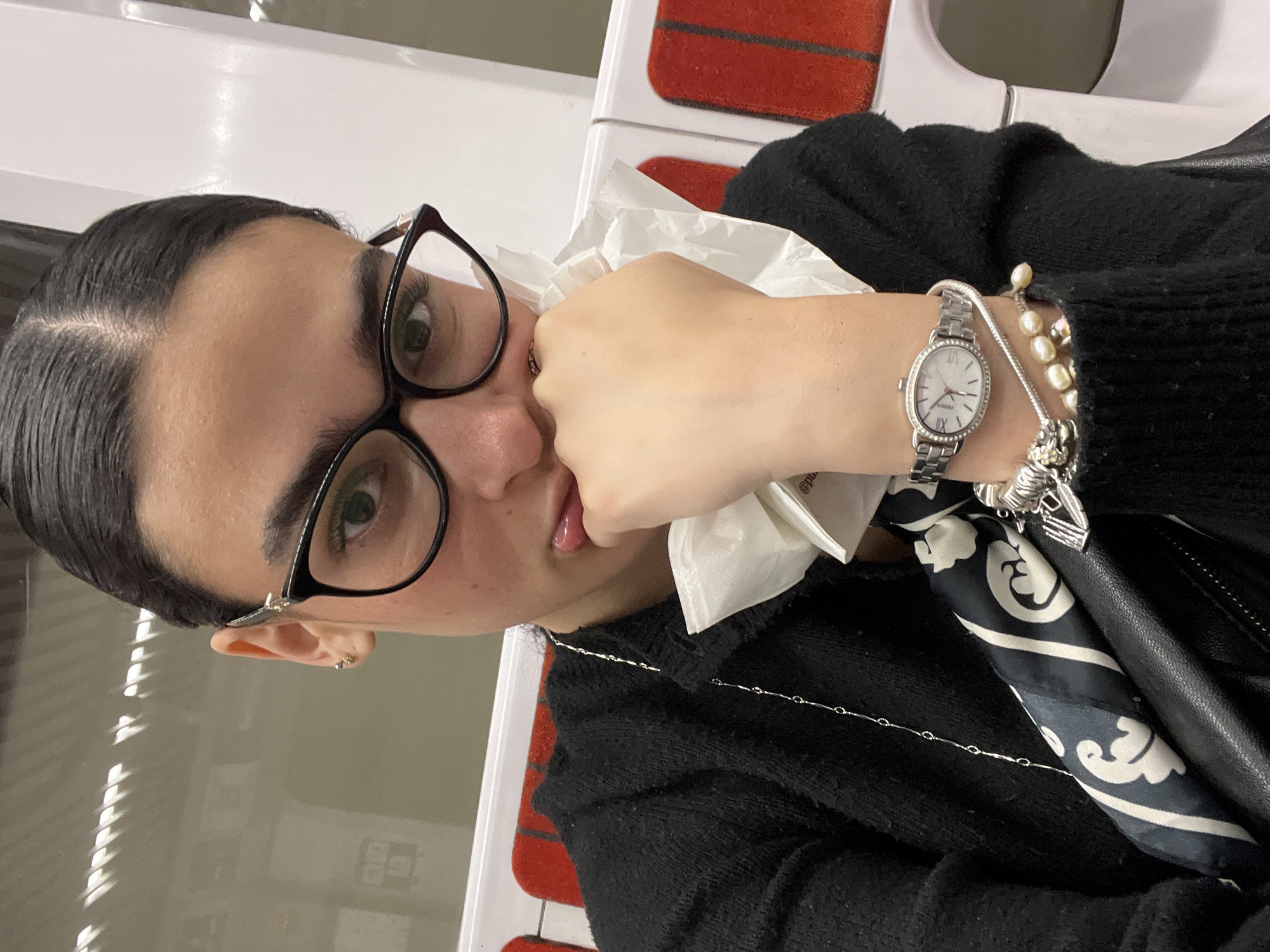 The image shows a person, likely female, from the chest up, positioned at an angle, looking directly at the camera. She has dark hair with a middle part, pulled back from her face, and wears black-framed glasses that highlight her green eyes. She is dressed in a black sweater.
On her left wrist, she wears a silver-toned watch with an oval face, Roman numerals, and the brand name "FOSSIL" visible. Alongside the watch are multiple bracelets, including one made of white pearls and another silver-toned chain bracelet with charms. A thin silver chain necklace is visible across her chest. Her left hand is raised to her mouth, holding a crumpled white tissue or napkin, partially obscuring her mouth which is slightly open, revealing a glimpse of her tongue. Partially visible text "@pio" can be seen on the tissue. A small gold-colored stud earring is visible in her left earlobe. Tucked near her right arm, a black and white patterned scarf is visible, possibly part of her outfit or accessories in a bag.
The setting appears to be indoors, possibly a public transport vehicle like a train or subway car, indicated by the white interior walls and segments of red seating visible in the background. The lighting is artificial. No specific time of day or weather can be determined. The person's expression is neutral.