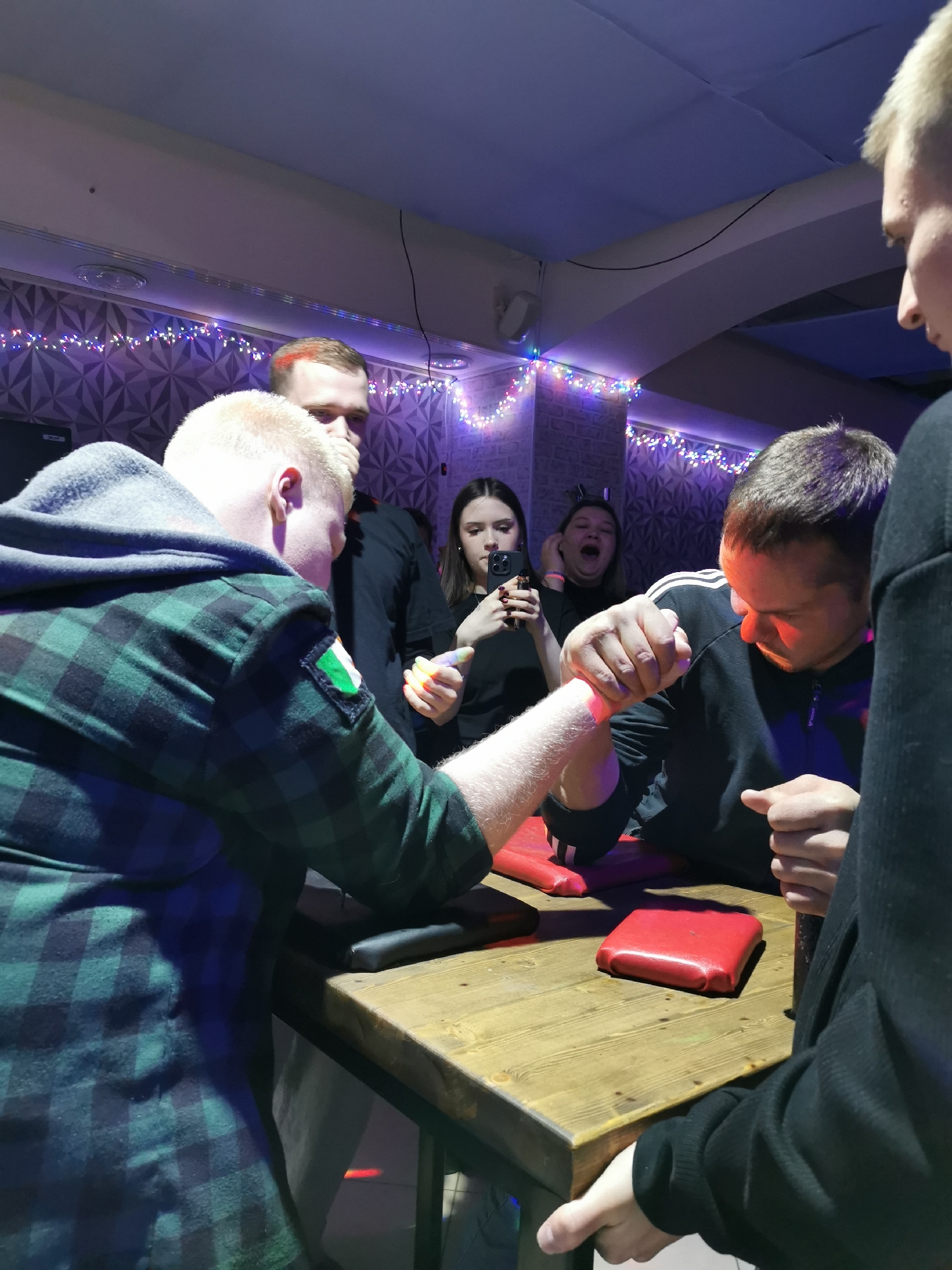 An indoor scene captures an intense arm wrestling match between two men. In the foreground, on the left, a man with light, short hair wears a green and black plaid shirt with a grey hooded sweatshirt underneath. An Irish flag patch (green, white, orange vertical stripes) is visible on his right arm sleeve. He grips the hand of his opponent across a wooden arm wrestling table. His opponent, on the right, has dark hair and wears a dark athletic jacket with white stripes on the sleeve. His face is illuminated with a reddish glow, reflecting the intensity of the competition. Both men's forearms also show a reddish hue, possibly from the red elbow pads on the table.
The wooden table has red upholstered pads for the competitors' elbows. Several onlookers surround the table. Behind the left arm wrestler, another man observes the match. Behind the right arm wrestler, a woman records the event on a smartphone, while another woman with her mouth open appears to be cheering or reacting vocally. A fourth person is partially visible on the far right.
The setting appears to be an evening event or party, indicated by the artificial lighting. Decorative string lights with small purple, pink, and white bulbs adorn the textured walls in the background, which feature a geometric pattern. The atmosphere is energetic and focused on the arm wrestling match. No discernible text is visible in the image.