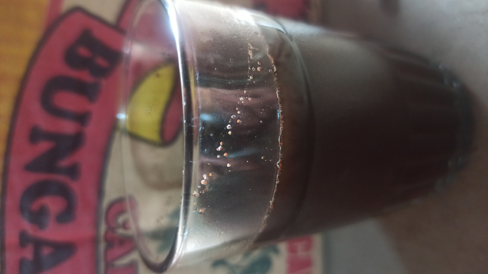 A close-up, slightly blurred image shows a clear glass filled with a dark liquid, likely coffee or tea, sitting on a neutral-colored surface. The liquid has a thin layer of foam or sediment on top, with small particles visible on the interior surface of the glass above the liquid line. In the background, a partially visible sign with red and yellow lettering in a foreign language, possibly Indonesian, is out of focus. The overall impression is of a casual, indoor setting.
