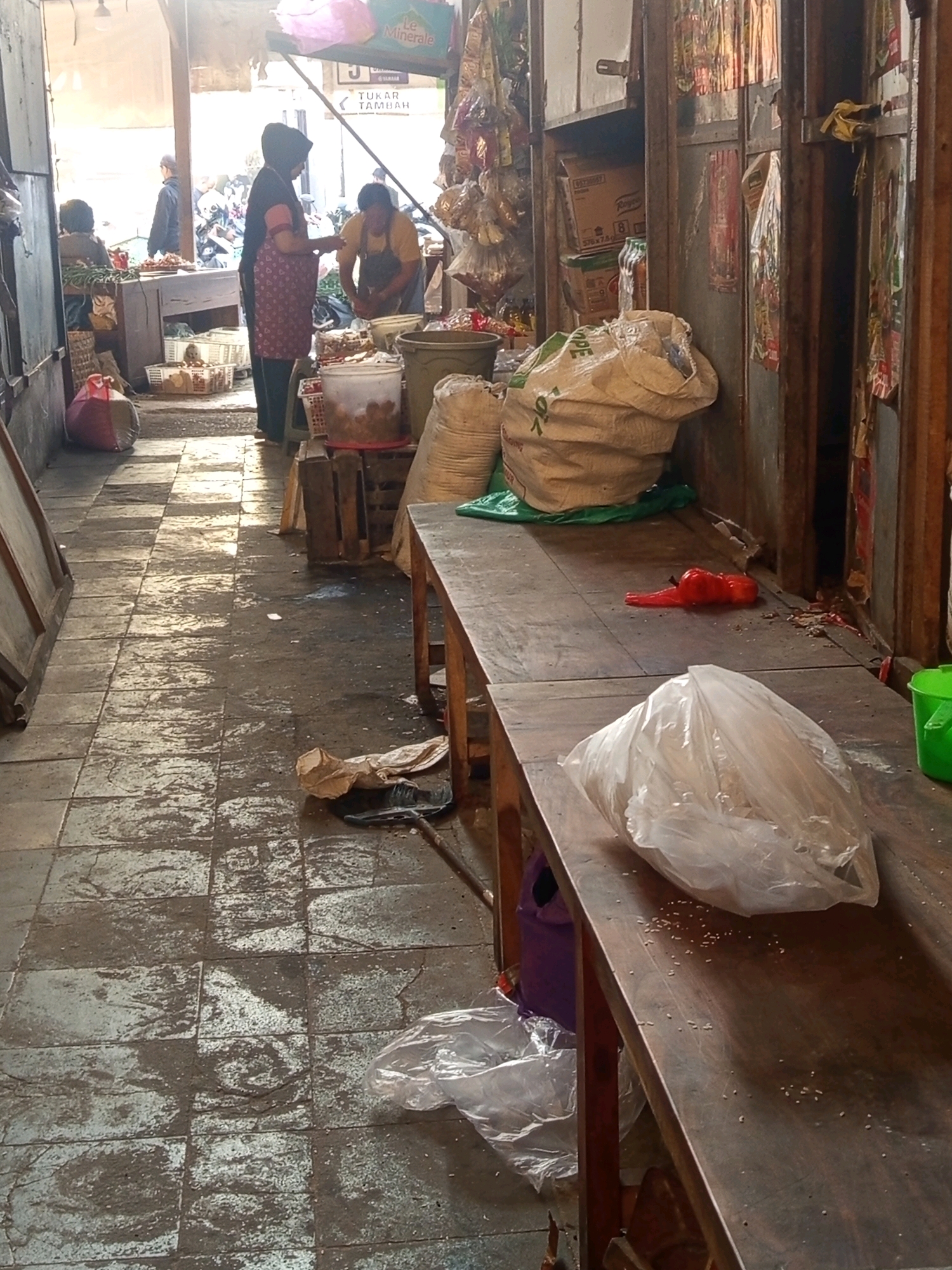 The image depicts a market scene in Tuban, Indonesia. In the background, a woman in traditional attire is handing something to a person wearing an apron, likely a vendor. Other figures are visible further back near motorcycles. The foreground features a long wooden table with various items, including a large white sack, a plastic bag filled with a white substance, a red object, and scattered grains. The floor is tiled and appears wet and dirty, suggesting a busy, possibly outdoor, market environment. The overall impression is one of everyday commerce in a local Indonesian setting.