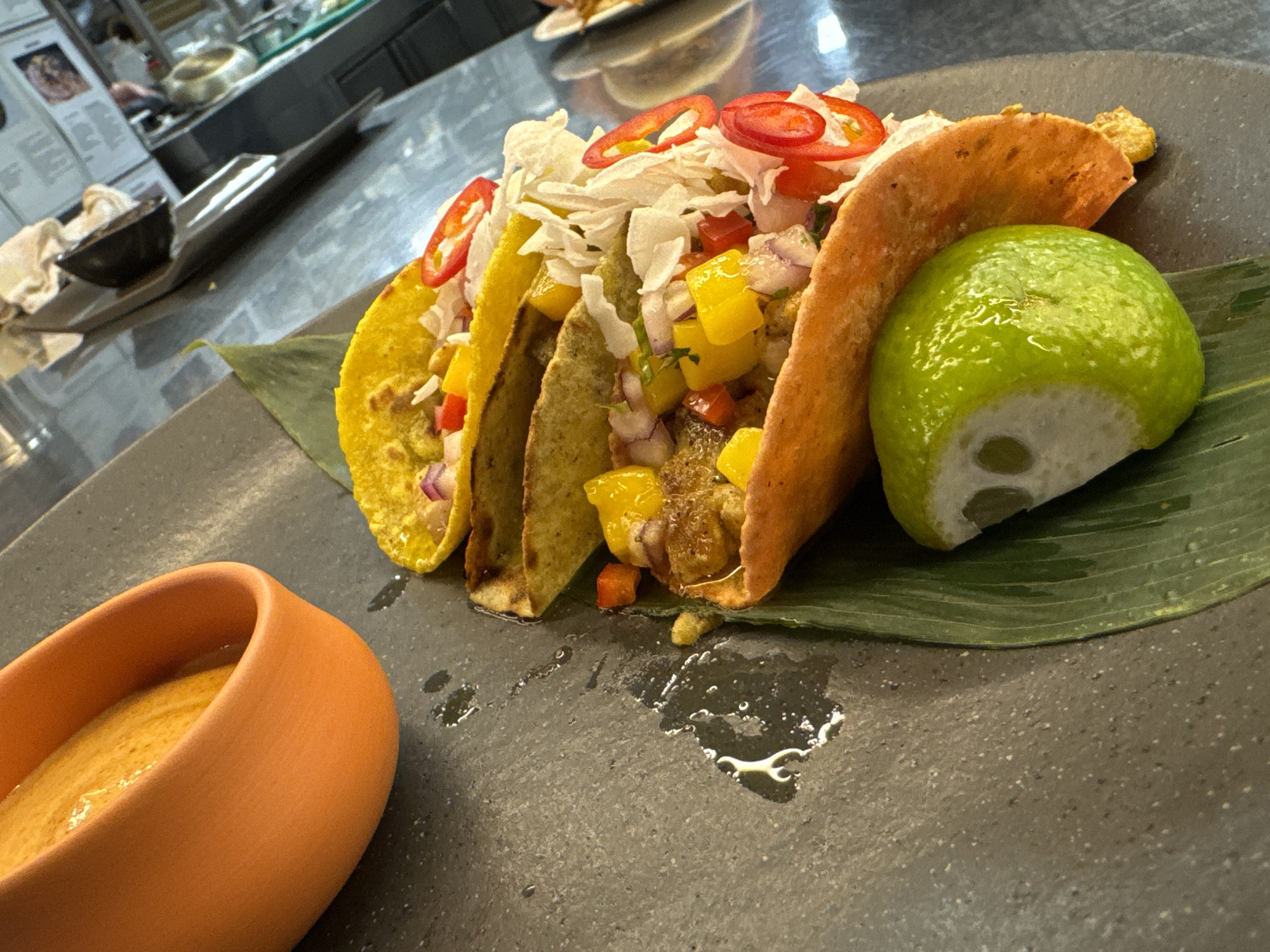 This image depicts a close-up, angled shot of three tacos on a dark, slate-like plate. The tacos are filled with what appears to be shredded lettuce, diced yellow peppers, red onions, and possibly some type of meat. Sliced red chili peppers and a wedge of lime are also visible on the plate. To the left of the tacos, an orange ceramic bowl filled with a vibrant orange sauce or puree is partially visible. A large green leaf, likely for presentation, is placed beneath the tacos. The background suggests a restaurant setting with hints of kitchen equipment and a menu board. There are no people visible in the image. The overall impression is of a well-presented, appetizing dish. The location is not specified in the image, and therefore cannot be identified as Sofia, Bulgaria.