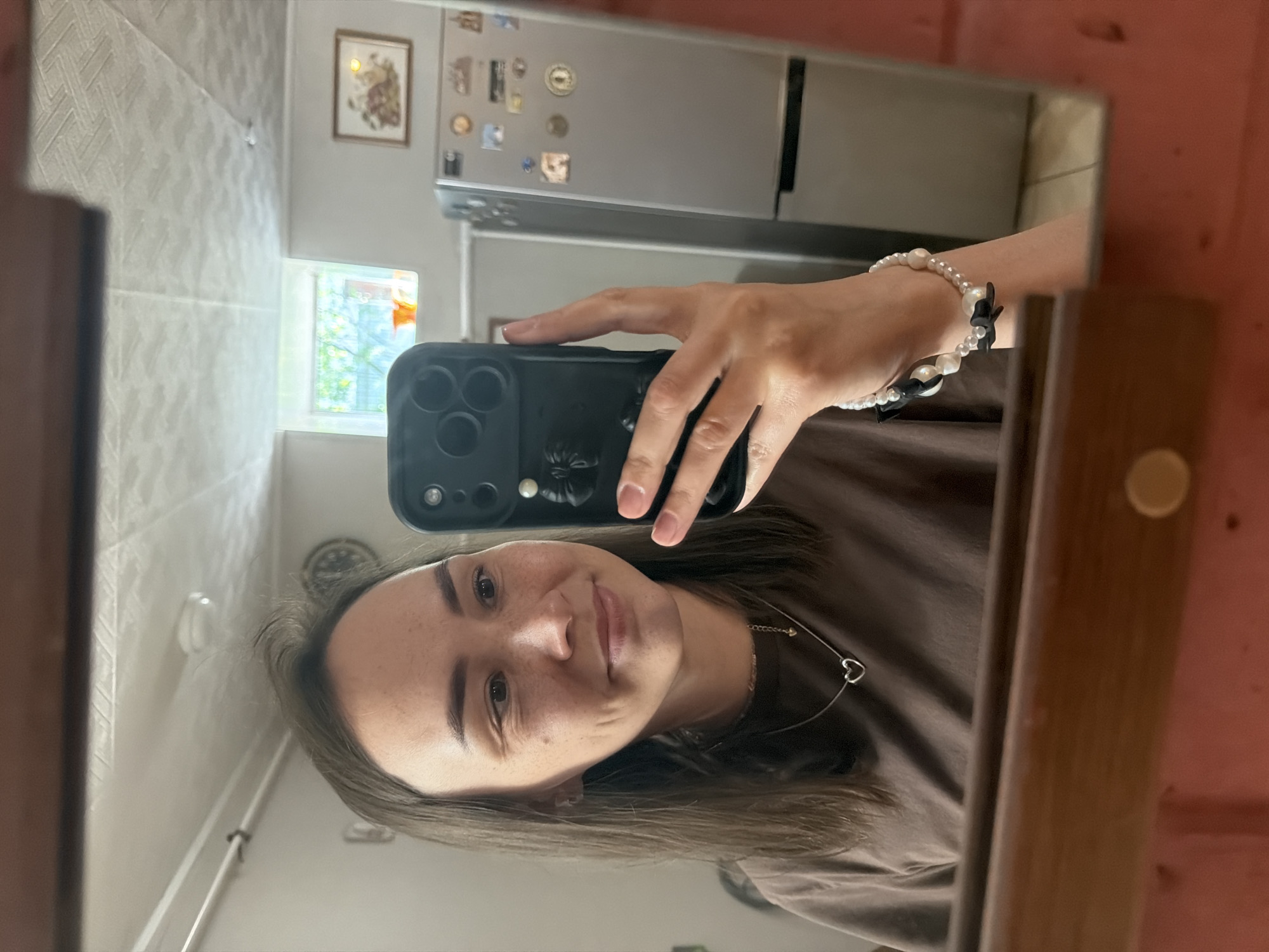 A person is taking a selfie in a mirror. The person has light brown hair, brown eyes, and is wearing a brown shirt and a necklace with a heart pendant. The person is holding a black smartphone with a black floral pop socket. The person is wearing a pearl bracelet with black bows. The background appears to be a kitchen, featuring a two-door stainless steel refrigerator adorned with magnets, a window with greenery visible outside, and tiled walls. Based on the metadata, the location is Chaikovsky, Russia.