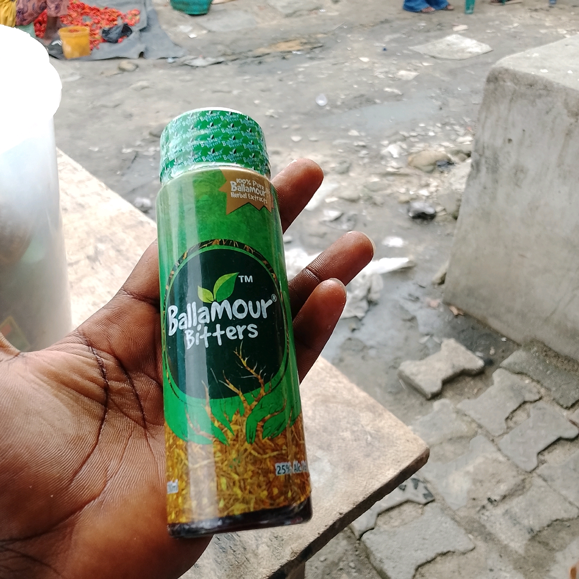 The image shows a close-up of a person's hand holding a green bottle of "BallAmOur Bitters Herbal Extracts". The bottle has a green, patterned cap and a label with the product name and "100% Pure" printed on it. The background appears to be an outdoor setting with a rough, grey surface, possibly a street or market area, with some scattered debris and a concrete block visible on the right. The overall lighting suggests it is daytime. The context of Ado, Nigeria is not directly evident from the visual information in the image itself.