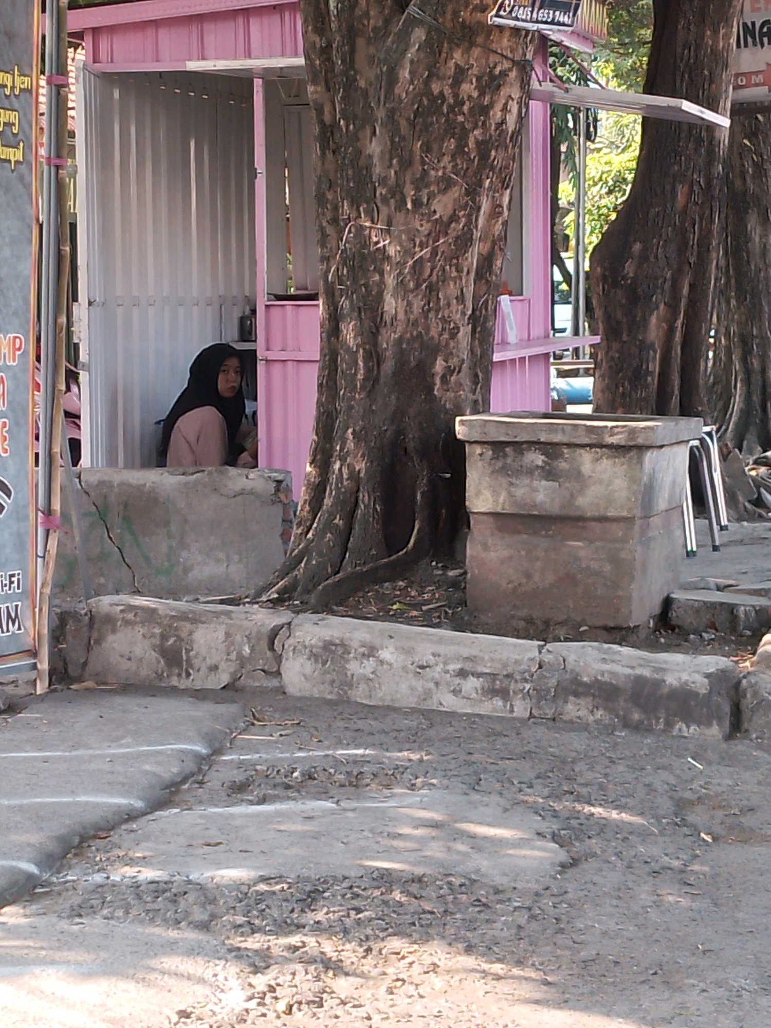 The image depicts a street scene in Kertosono, Indonesia. In the background, a pink food stall is visible, partially obscured by trees. A woman wearing a black hijab and a beige top is seated inside the stall, looking towards the camera. In the foreground, a concrete structure, possibly a planter or a bench, is positioned on the side of the road. The road surface is asphalt with visible cracks and patches. Several large trees with rough bark line the street. The overall environment suggests an outdoor setting with some urban elements.