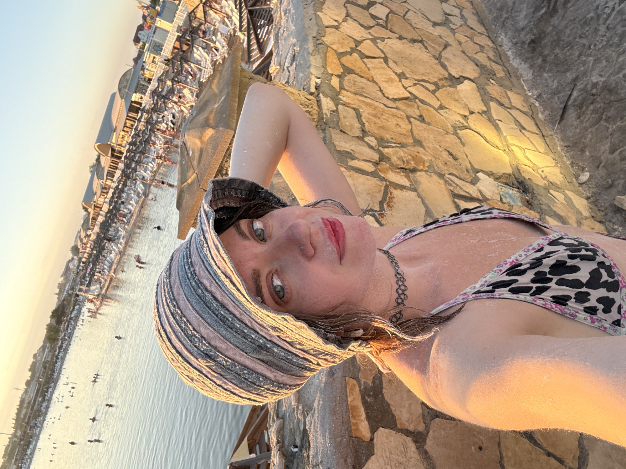 A woman in a bikini and a bucket hat poses for a selfie against a backdrop of a sunlit lake and resort area. The woman's face is partially obscured by her hat, but her green eyes are visible. She has on a patterned bikini top and a choker necklace. Her skin appears to be covered in salt, suggesting she is at a salt lake or spa. In the background, people are swimming in the lake, and lounge chairs and umbrellas are set up on the shore. The scene is bathed in the warm glow of sunset, casting long shadows and illuminating the water. The location is the salt lake of Sol-Iletsk, Russia, known for its therapeutic properties.