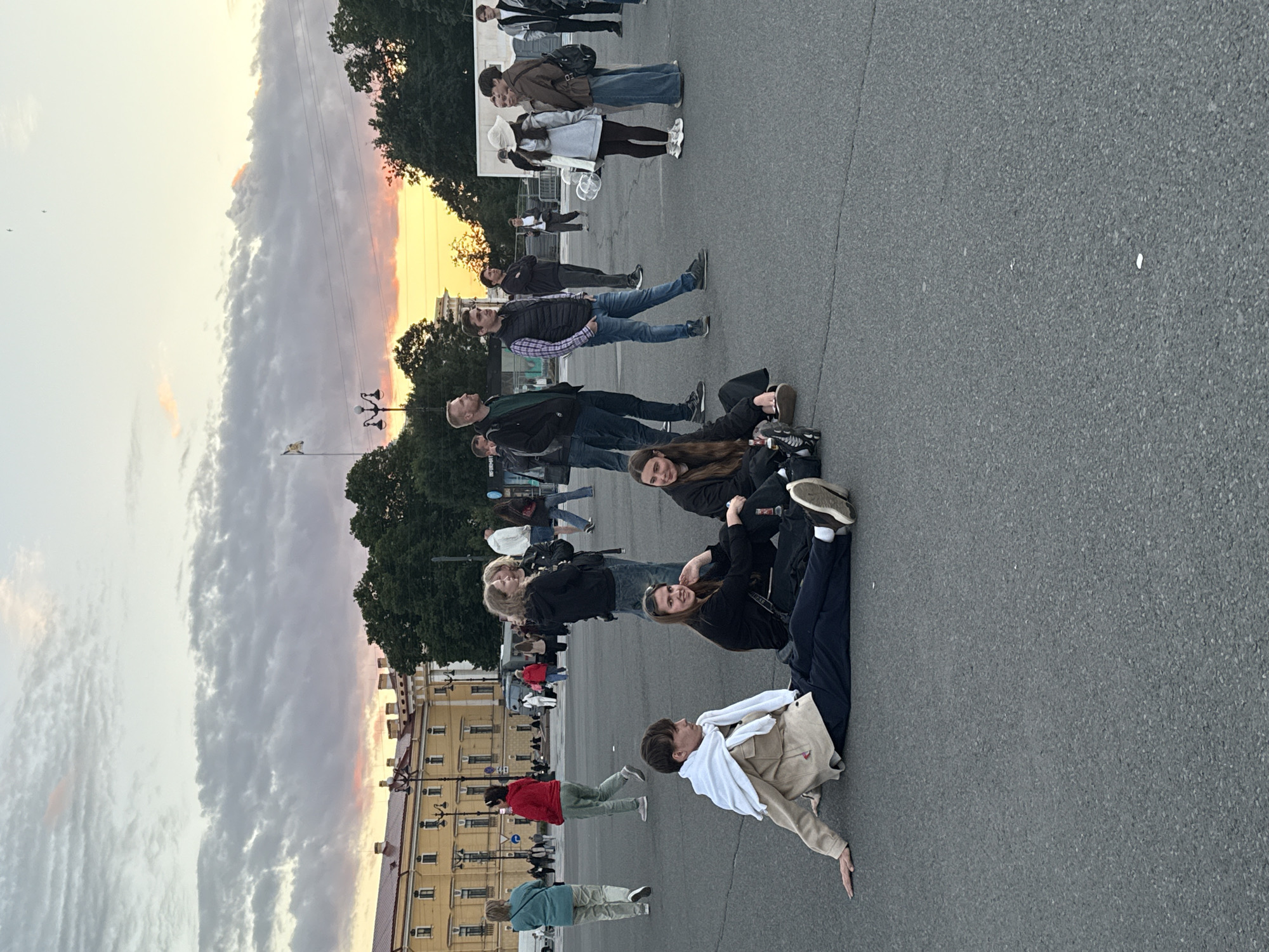 The image depicts a wide-angle view of a large paved public space in Saint Petersburg, Russia, likely Palace Square, during what appears to be a late afternoon or early evening, judging by the sunset sky. A diverse group of people are scattered across the area, some standing and others seated on the ground.
In the foreground, four young individuals are seated on the pavement. One male, wearing a light-colored jacket draped over his shoulders and dark trousers, is reclined on his left side with his right arm extended. Next to him, a female in a black top and dark trousers is sitting with her legs crossed, looking towards the camera with a slight smile. Another female, also in a black top and dark trousers, sits beside her, appearing to be engaged in conversation.
Further back, a larger gathering of people are seen. Men in casual attire, including jeans and jackets, are visible, some conversing, others looking around. Several women are also present, some in jeans and tops, one in a light-colored dress and hat, and another in a brown jacket and jeans. The background features a large, classical yellow building with a red-tiled roof, characteristic of Saint Petersburg's architecture. Trees line the perimeter of the square, and a flag is visible on a pole. The sky above is dramatic with clouds, colored by the setting sun, creating a warm orange and pink glow. The overall atmosphere appears relaxed and social.