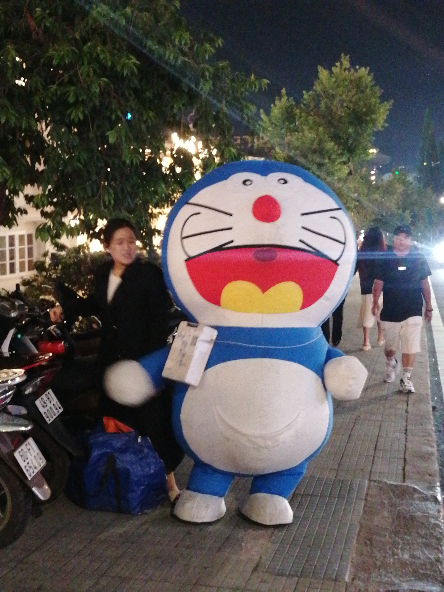The image depicts a person in a Doraemon costume on a sidewalk in Da Lat, Vietnam. A woman in a black jacket is near parked motorbikes. Several people are walking on the sidewalk behind the Doraemon character. The lighting suggests it is evening. Trees and buildings are visible in the background, indicative of an urban setting.