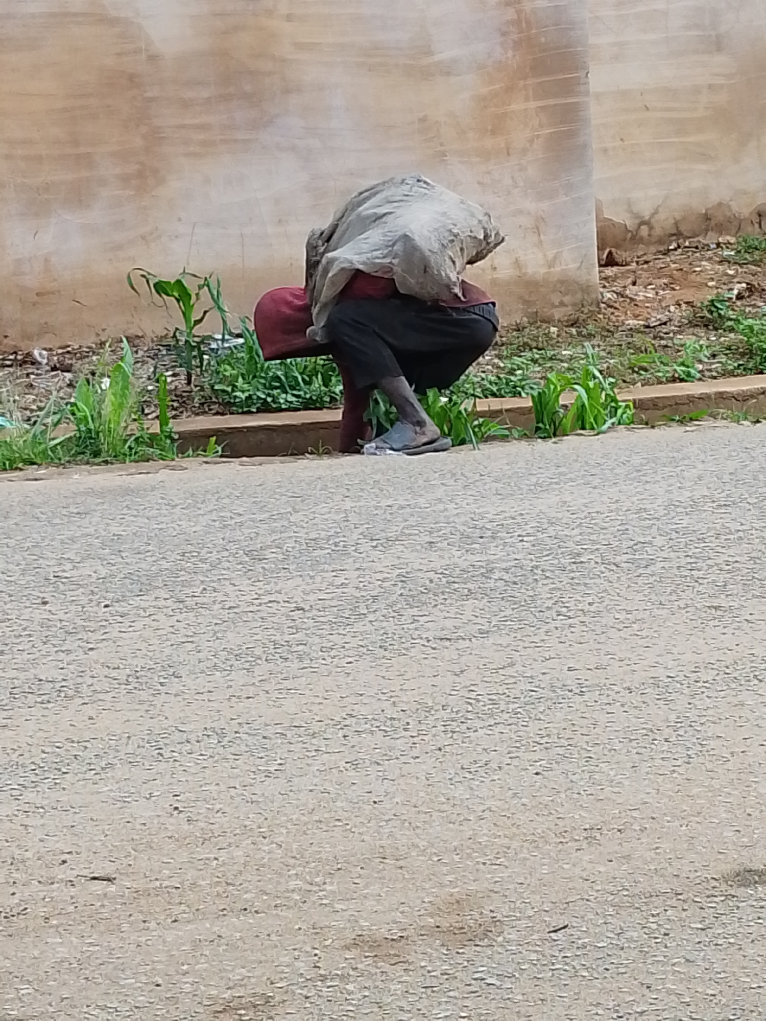 The image shows a person sitting on a red stool in Jos, Nigeria. The person is draped in a gray cloth, obscuring their head and upper body. They are wearing black pants and a gray sandal on one visible foot. The person is positioned on a narrow strip of land between a paved road and a concrete wall with vegetation growing along its base. The road surface is gray and textured. Behind the wall is a patch of brown dirt with scattered debris and additional vegetation.