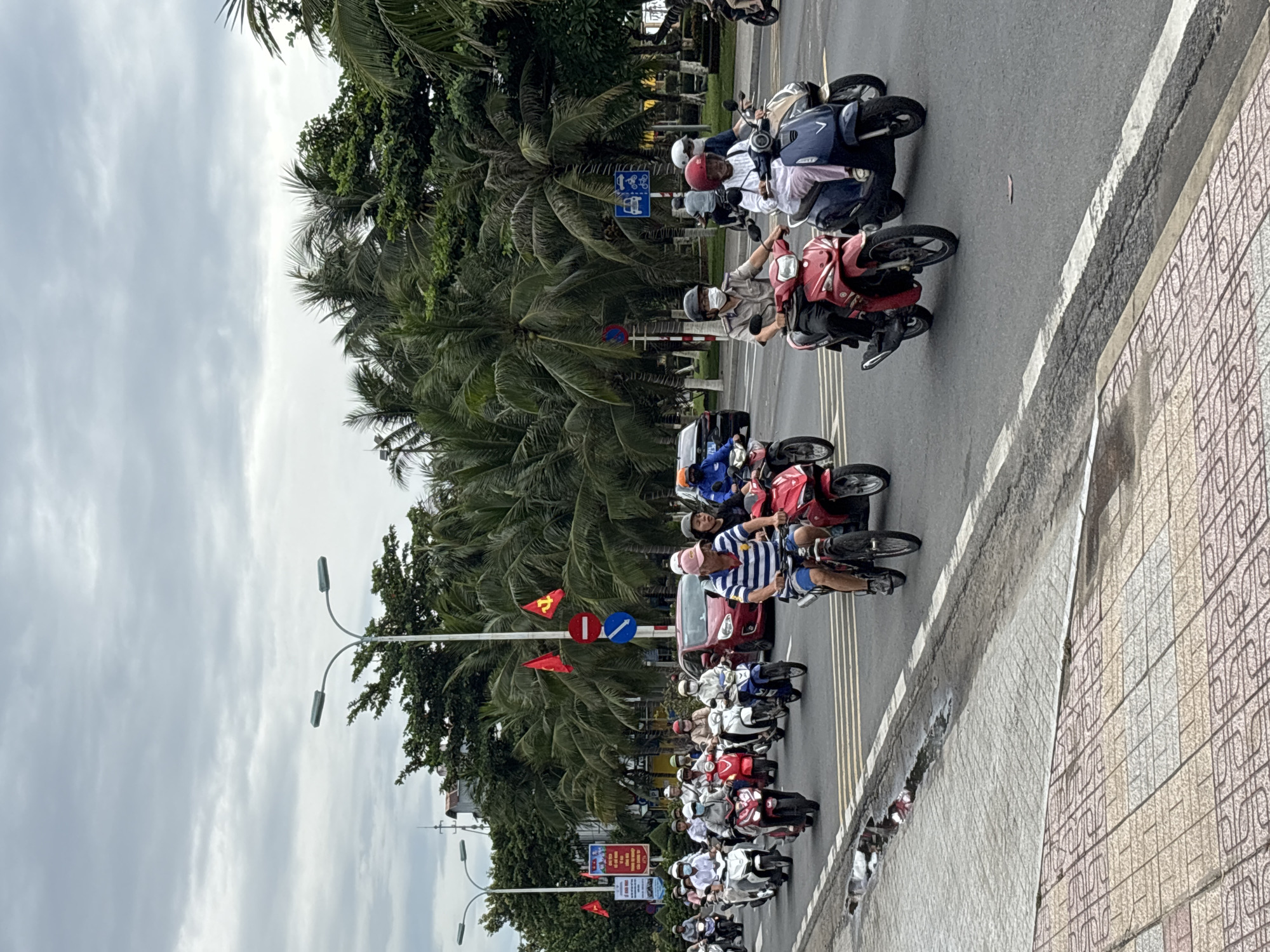 A group of people are riding motorcycles on a street in Nha Trang, Vietnam. The sky is overcast, and palm trees line the street. The motorcycles are of various colors, including red, white, and blue. Some of the riders are wearing helmets, and some are wearing masks. There are also a few cars on the street. In the background, there are buildings and signs. The street is paved with asphalt, and there are white lines painted on the road to indicate lanes. The sidewalks are paved with patterned tiles.