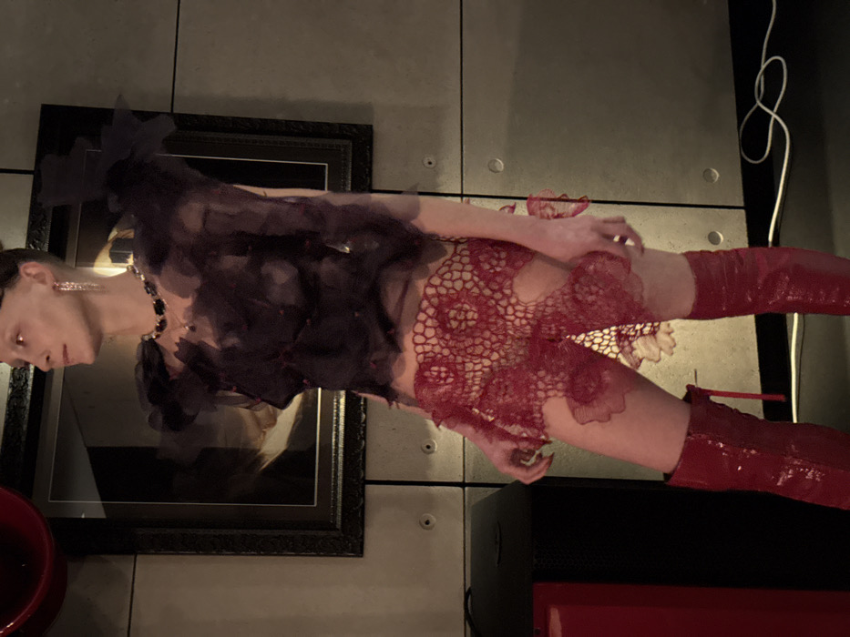 A slender individual is depicted standing indoors, facing slightly right with their head turned further right, in what appears to be a dimly lit, contemporary interior. The person is dressed in a dark, semi-sheer top featuring ruffled or layered fabric with scattered red embellishments, an intricate red openwork lace-like mini-skirt, and shiny red knee-high boots. They wear long, dangling earrings and a dark beaded necklace; their hair is styled back, and eyebrows appear light. The background consists of dark grey or metallic paneled walls with visible seams and circular fastenings. A large, dark-framed picture is mounted on the wall behind the individual's upper torso. The dark floor exhibits a coiled white cable to the right, and a dark rectangular object is partially visible to the left. A fragment of a red, possibly reflective, object is present in the upper left corner. The specific location within the city of London, United Kingdom, cannot be determined from the visual evidence.