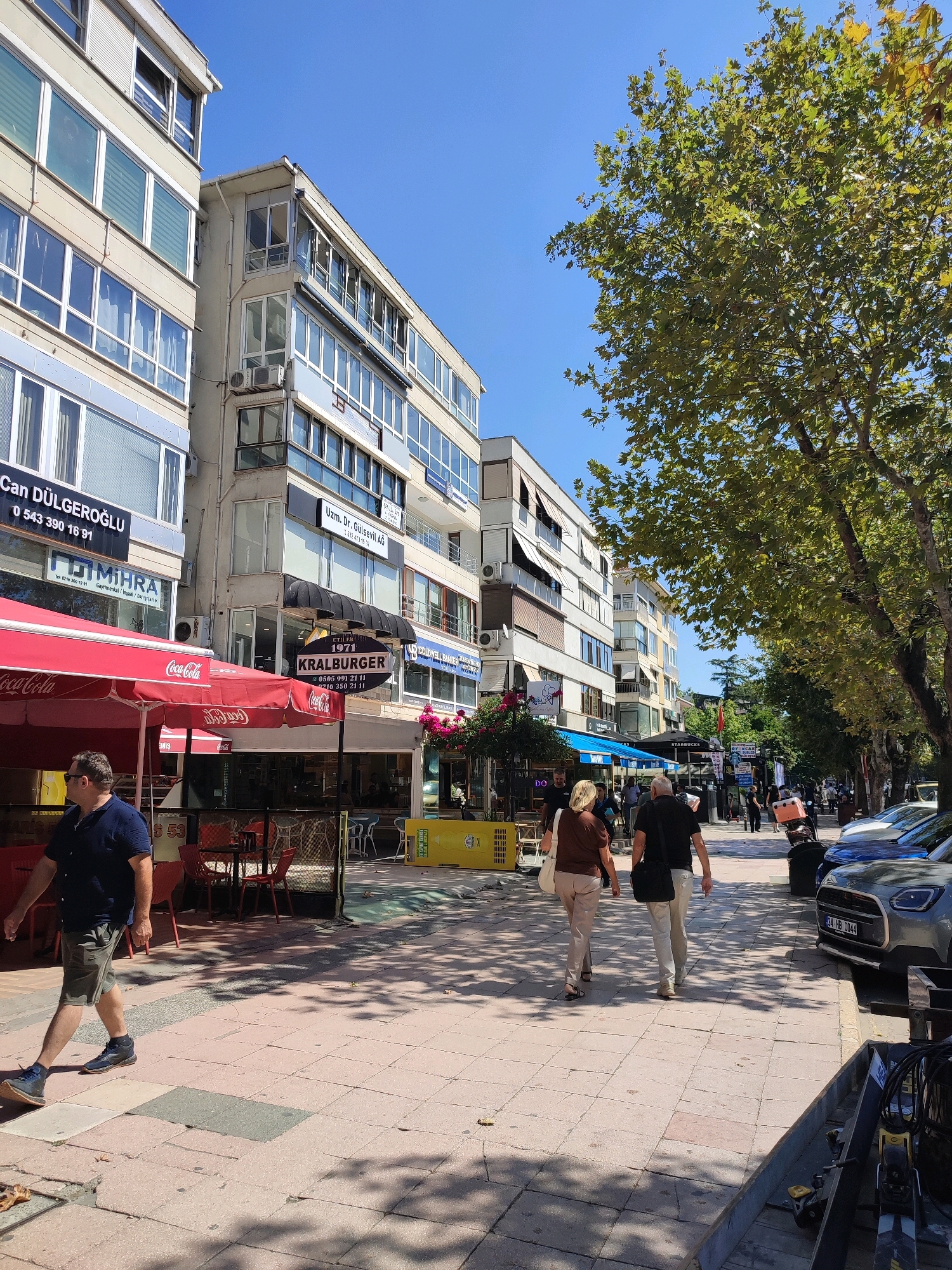 The image depicts a street scene in Kadıköy, Turkey, on a sunny day. In the foreground, a man wearing a navy blue short-sleeved shirt and grey shorts walks on a tiled sidewalk. Behind him, red and white umbrellas of a cafe are visible, along with tables and chairs. The buildings lining the street are multi-story structures with numerous windows. Signs on the buildings include "Can DÜLGEROĞLU," "FUMIHRA," "Uzm. Dr. Gülsevil AG," and "KRALBURGER." Further down the street, several people are walking, and cars are parked along the curb. A prominent tree with lush green foliage casts shadows on the sidewalk. The sky is clear and blue.