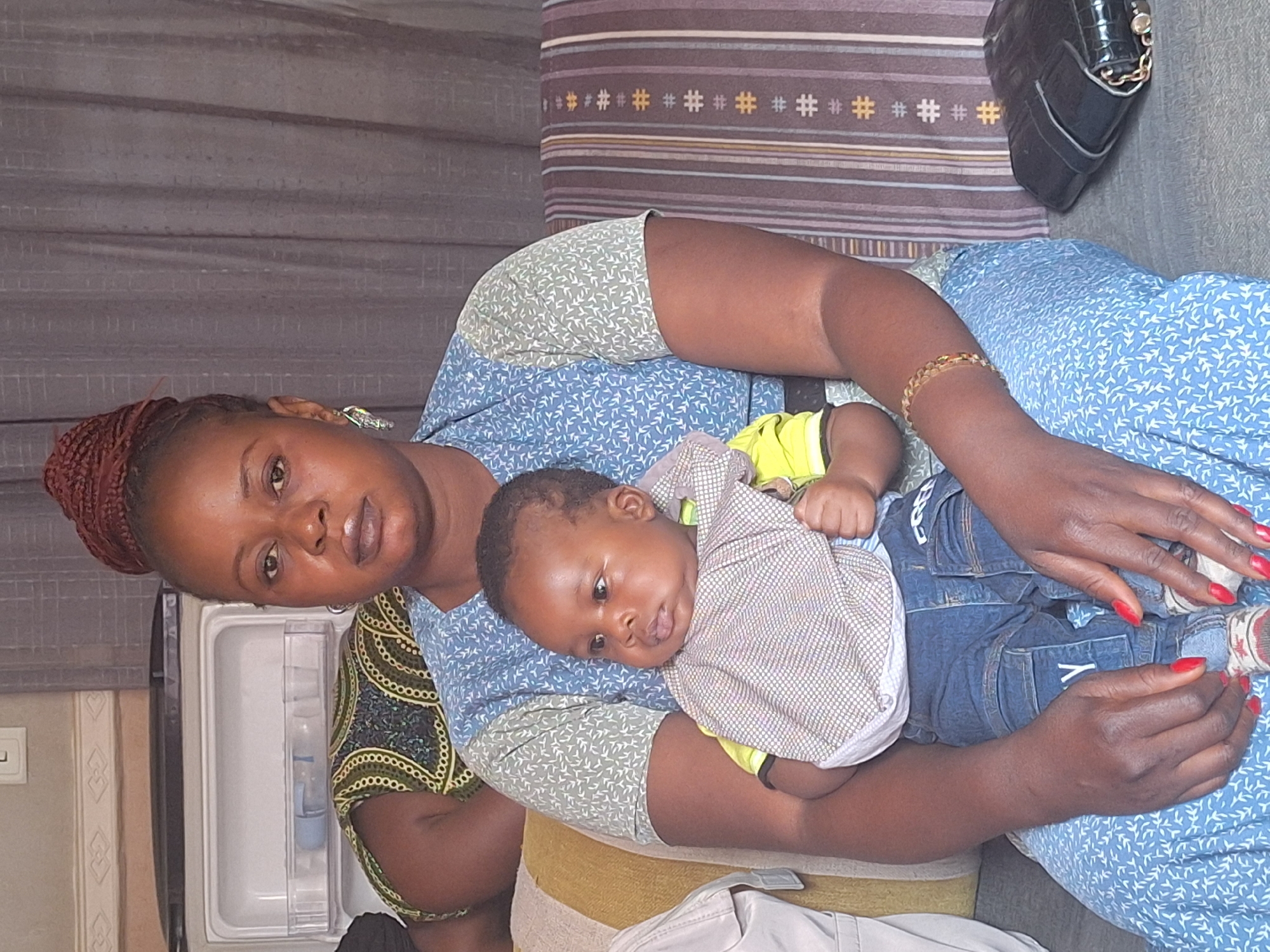 A medium shot shows a woman in a blue dress with a light green patterned top, seated and holding a baby. The baby is wearing a yellow shirt, a patterned bib, and blue denim overalls. The woman has dark skin, dark hair tied up on her head, and is wearing a gold bracelet. She is looking directly at the camera. A black purse sits on the sofa behind her. The photo was taken indoors in Sabon Tasha, Nigeria. In the background, there is a gray curtain, a striped pillow, and a white refrigerator.