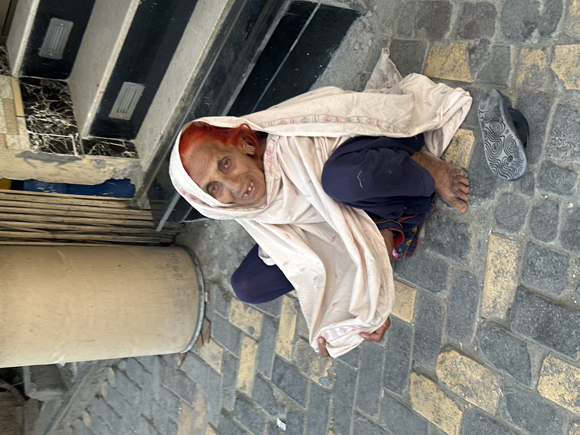 A photograph depicts an elderly woman squatting on a paved surface. She has red hair, her face is weathered with wrinkles, and she is looking upwards. She is wearing dark blue pants and a light-colored shawl with a patterned border is draped over her head and body. Her bare feet are visible on the ground near a black sandal with a patterned sole. To her left is a cylindrical object, possibly a drum, and behind her are stone steps. The location appears to be an urban area in Quetta City Tehsil, Pakistan, given the style of paving and building materials.