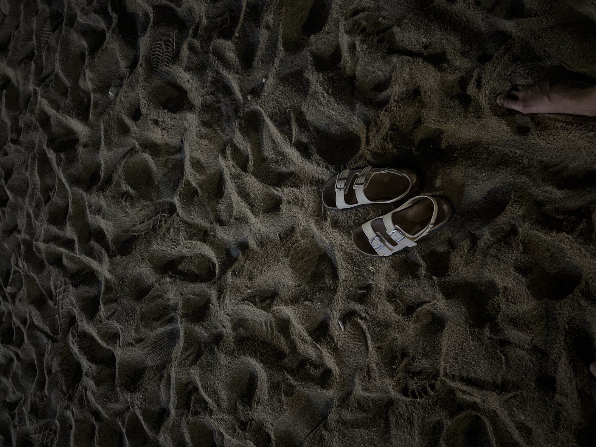 This media file presents an overhead view of dark, textured sand on a beach, likely during nighttime or under very low light conditions in Nha Trang Ward, Vietnam. The sand exhibits numerous ripples and indentations from various footprints, including distinct shoe prints, suggesting recent activity.
A prominent feature is a pair of light-colored sandals, possibly white or light grey with silver buckles, resting on the sand in the mid-right section of the frame. In the upper-right corner, a bare human foot is partially visible, hinting that the person has taken off their sandals and is standing or walking barefoot. The scene evokes a quiet, solitary atmosphere, characteristic of an evening or night stroll along the shoreline. Small fragments of debris are sparsely scattered across the sand. There is no visible text in the image.